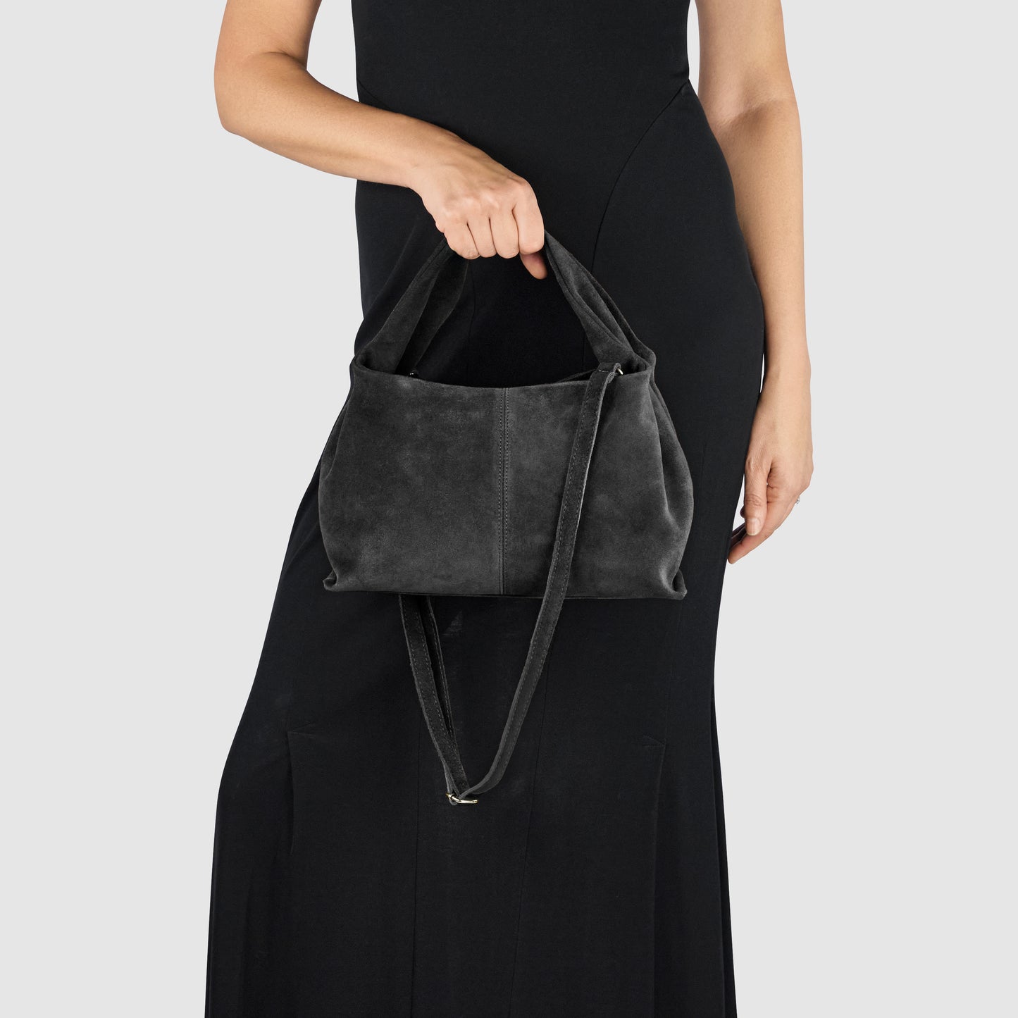 A person in a black dress holds a handcrafted bag made of black suede with a shoulder strap, standing against a plain light gray background.