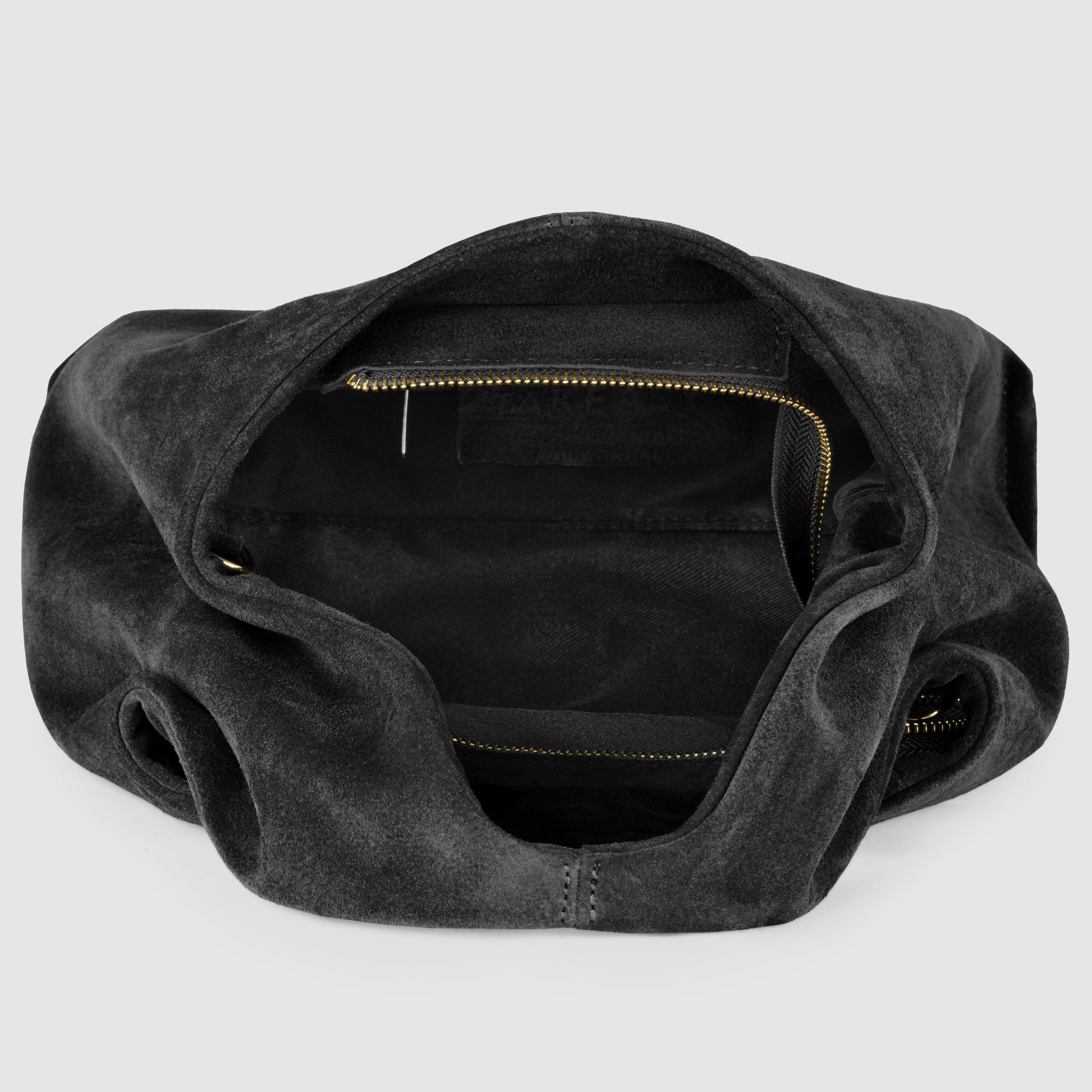 A top-down view of an open black suede handbag, showcasing Italian craftsmanship with a spacious interior, zippered pocket, and smooth lining. The empty bag is displayed against a light gray background.