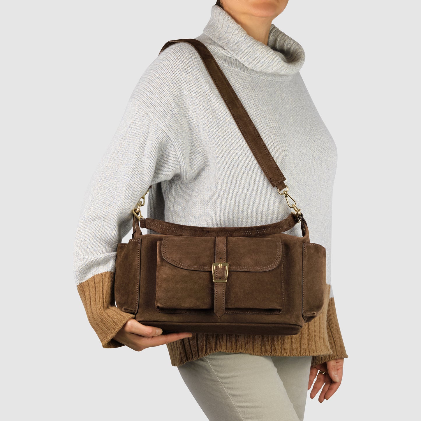 A person wearing a light gray and brown sweater and beige pants carries a handmade Italian handbag crafted from genuine suede leather, featuring a shoulder strap and front buckle pocket, against a plain background.