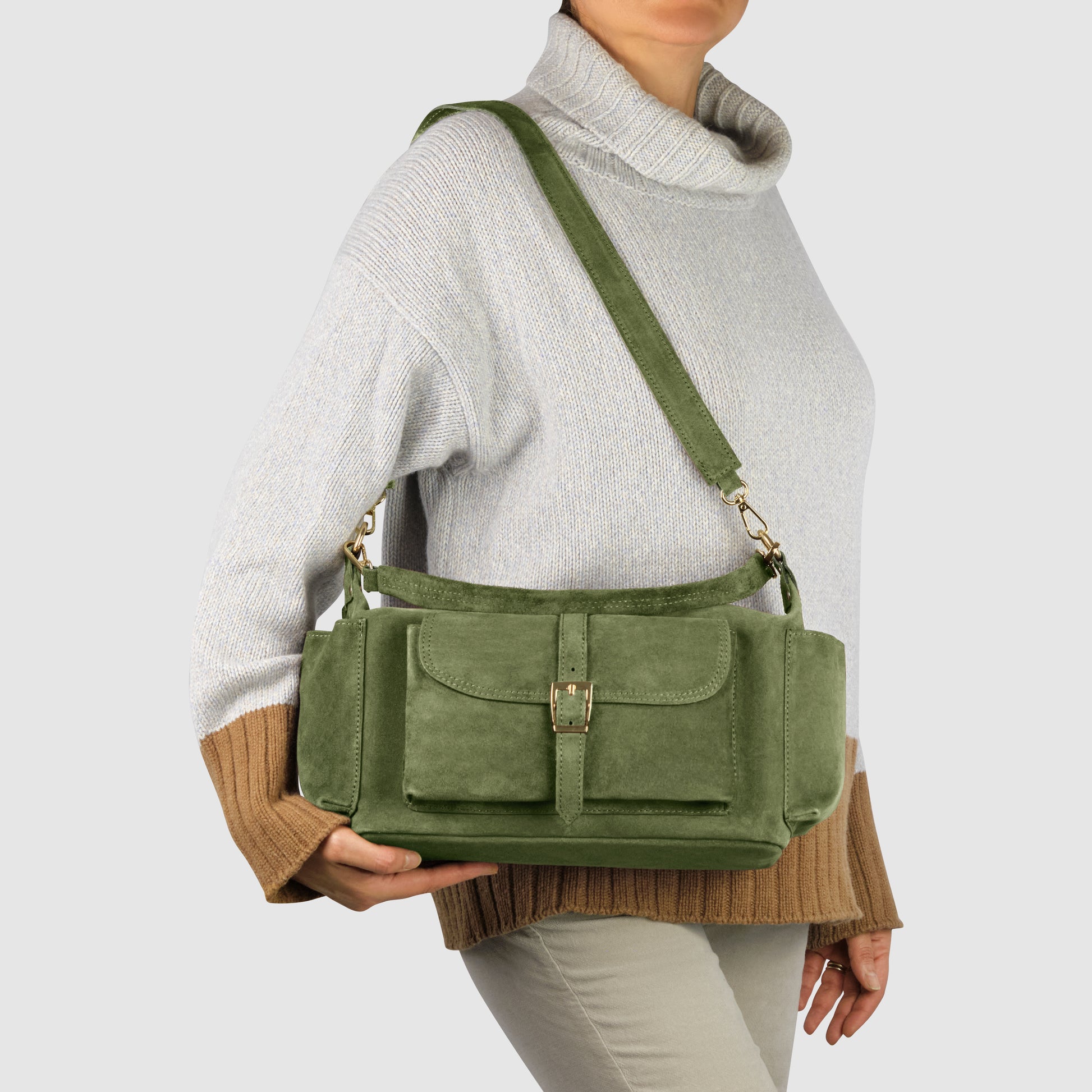 A person wearing a light gray and tan sweater holds a handmade Italian handbag crafted from genuine suede leather, featuring multiple pockets and a front buckle, carrying it by the strap over one shoulder against a plain background.