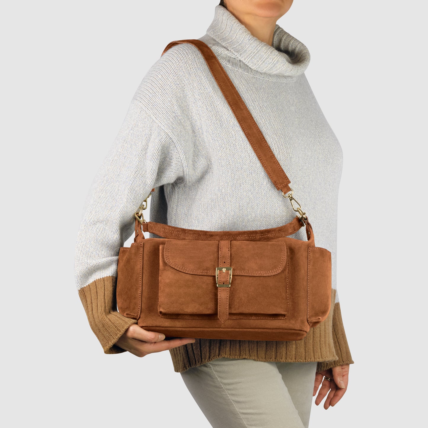 A person in a light gray turtleneck sweater and beige pants holds a handmade Italian handbag in brown genuine suede leather with front pockets and a buckle closure against a plain light background.