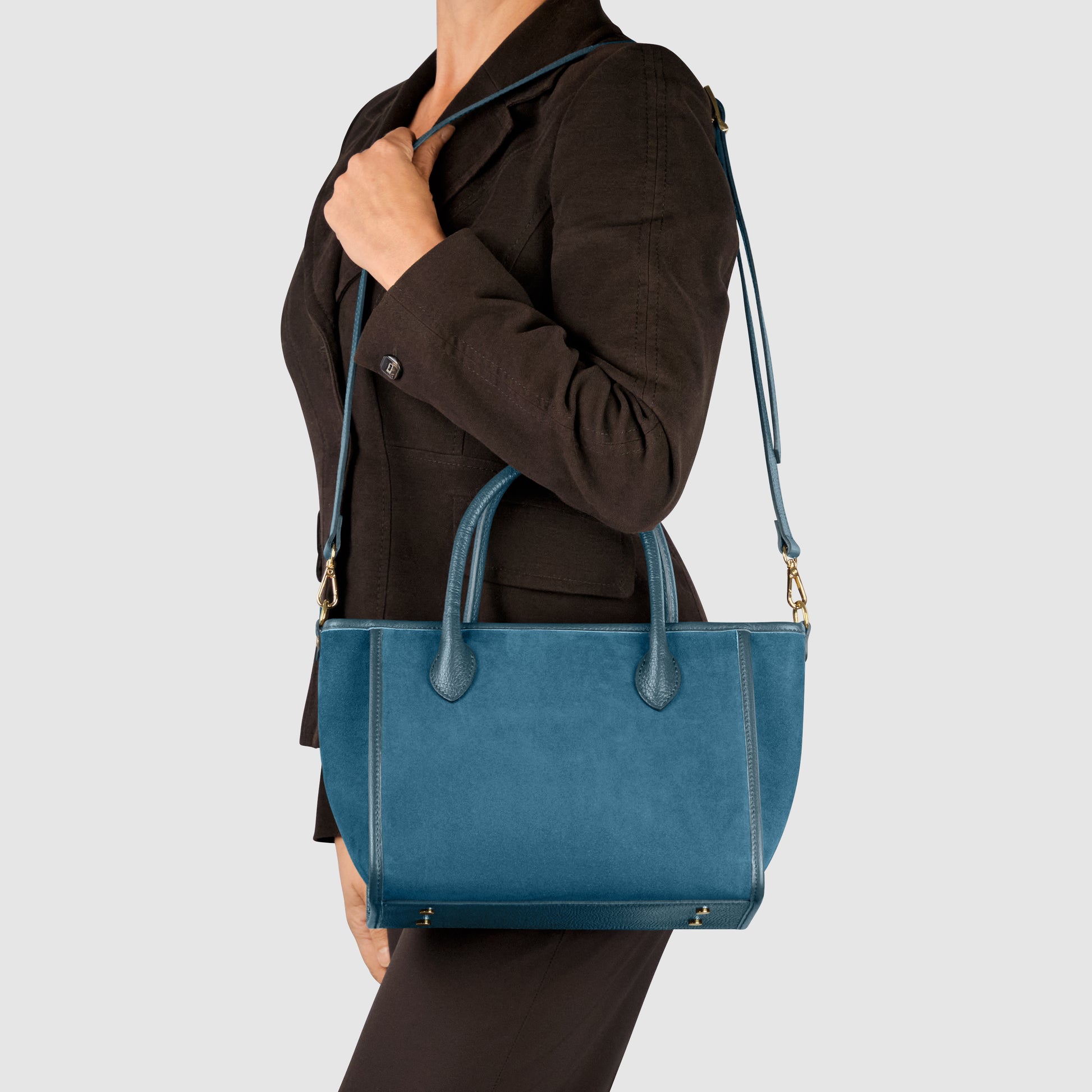 A person in a brown blazer and pants models an Everyday Elegance teal suede leather handbag, handcrafted in Florence, with both hand straps and a shoulder strap against a plain light gray background.