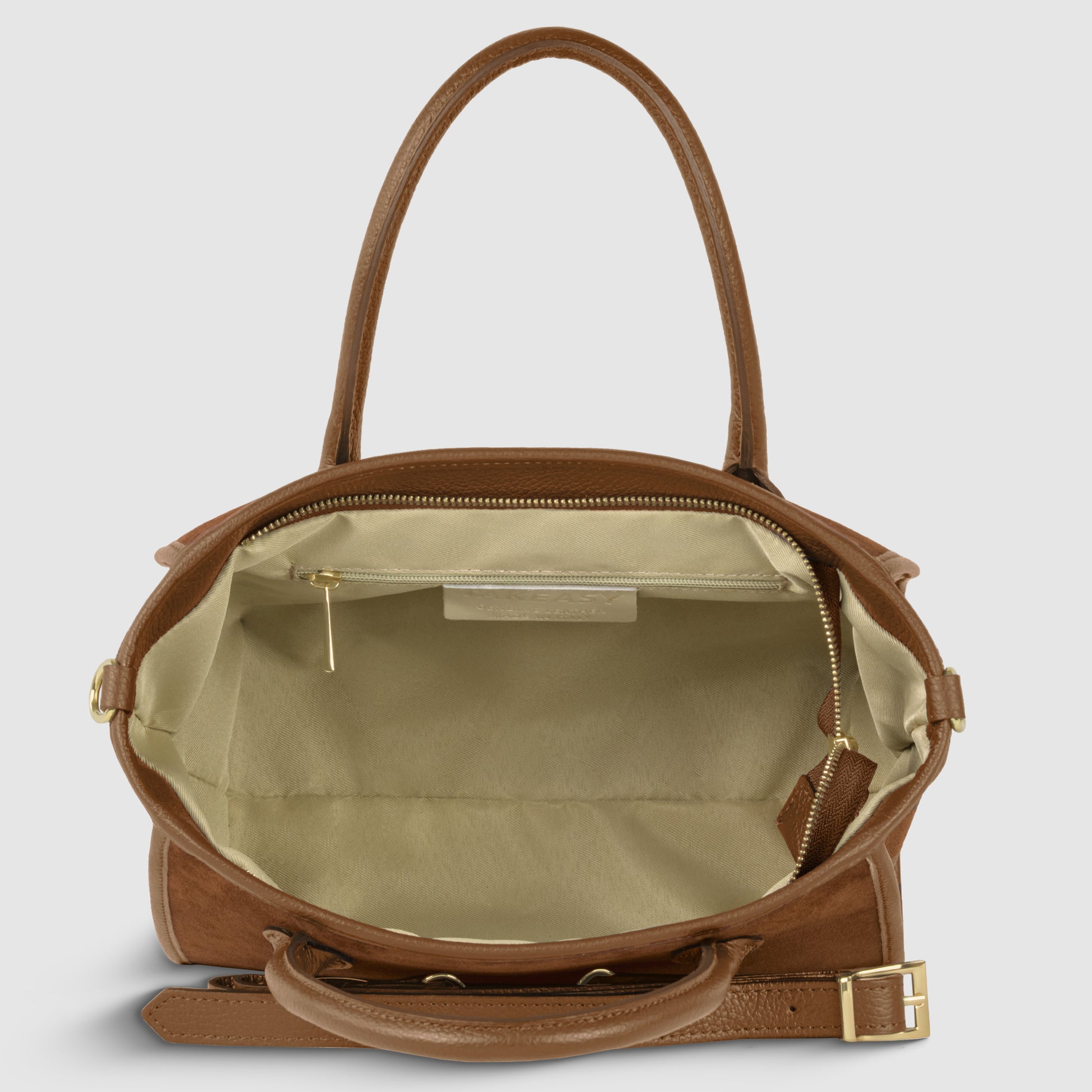 A brown suede leather handbag with gold hardware is shown open, revealing a spacious, light beige fabric-lined interior with a zippered pocket and small leather tag inside. The upright handles and adjustable strap complete this piece, handcrafted in Florence.
