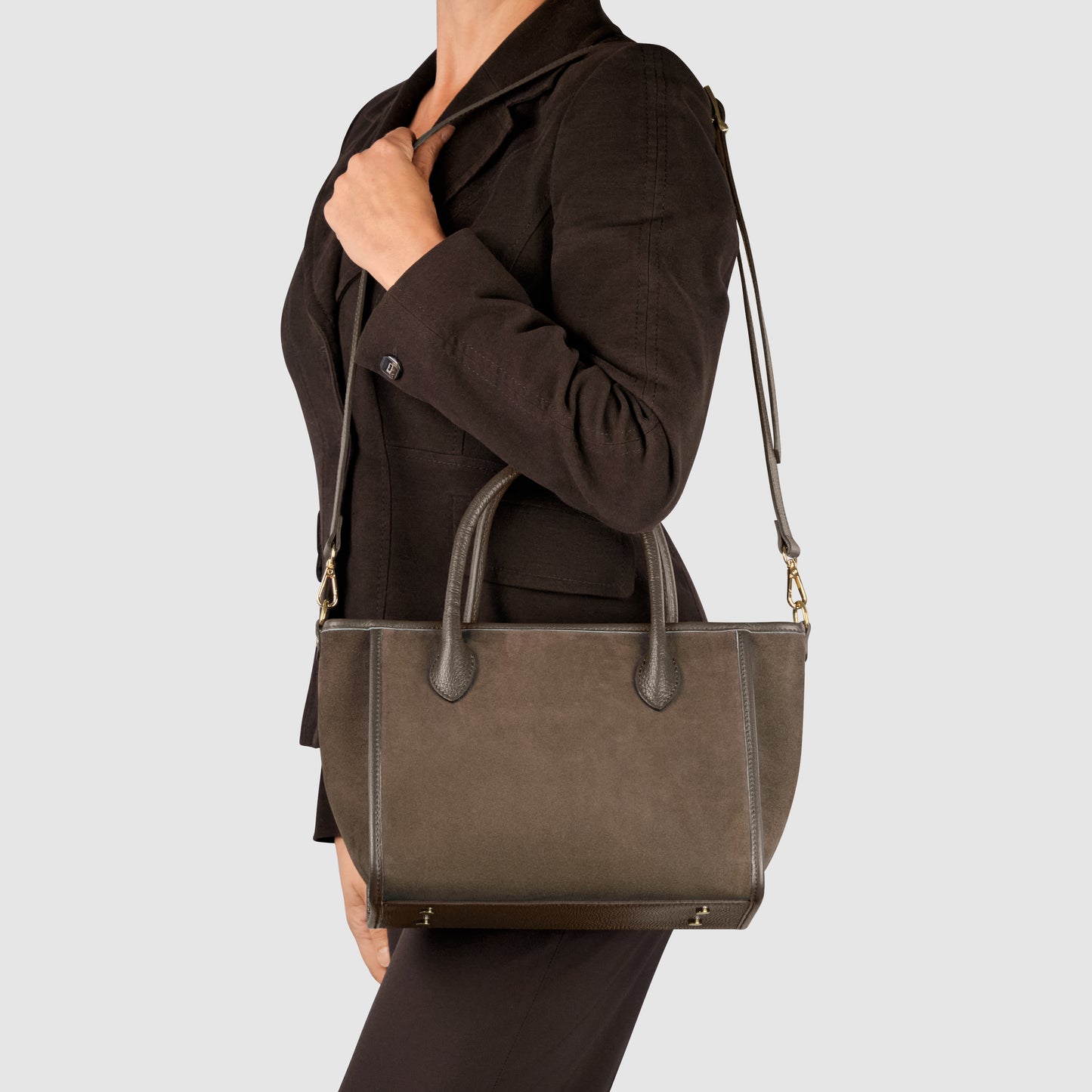 A person wearing a dark blazer holds a suede leather handbag with top handles and an adjustable detachable strap, shown from the side against a plain light background.