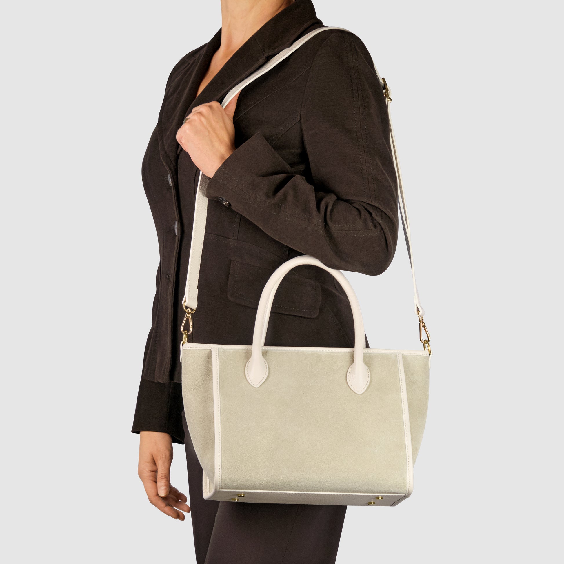 A woman in a dark blazer and pants carries a handcrafted in Florence suede leather handbag with both handles and an adjustable crossbody strap, shown against a plain light background.