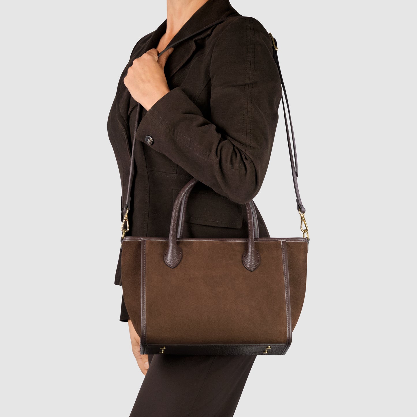 A person in a brown blazer and pants carries a handcrafted in Florence suede leather handbag with an adjustable detachable strap on their shoulder, shown in side profile against a plain light gray background.