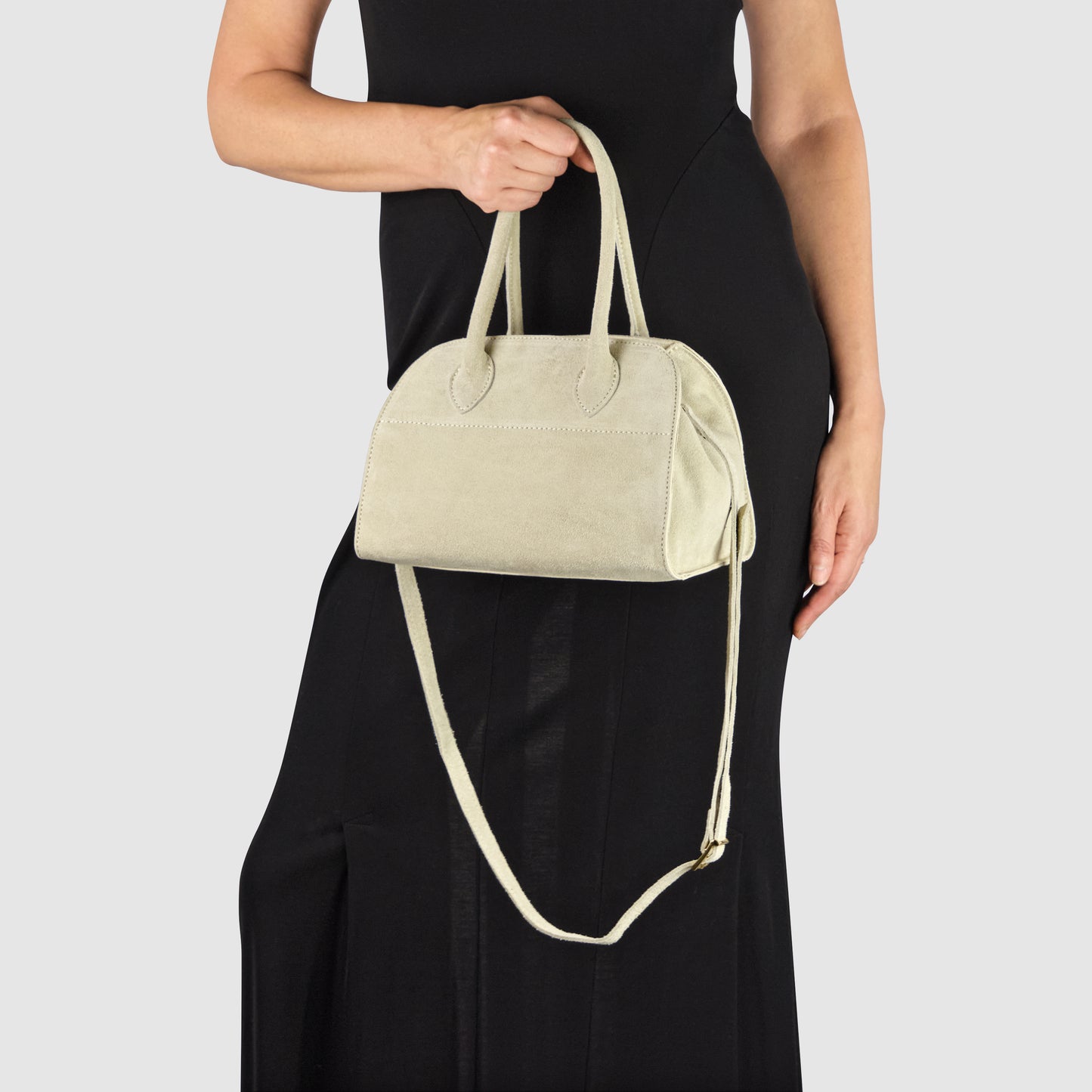 A person in a sleeveless black dress holds a handmade Italian handbag with both handles in one hand. The bag, crafted from Florentine leather, also features a long shoulder strap and a simple design. The background is plain light gray.