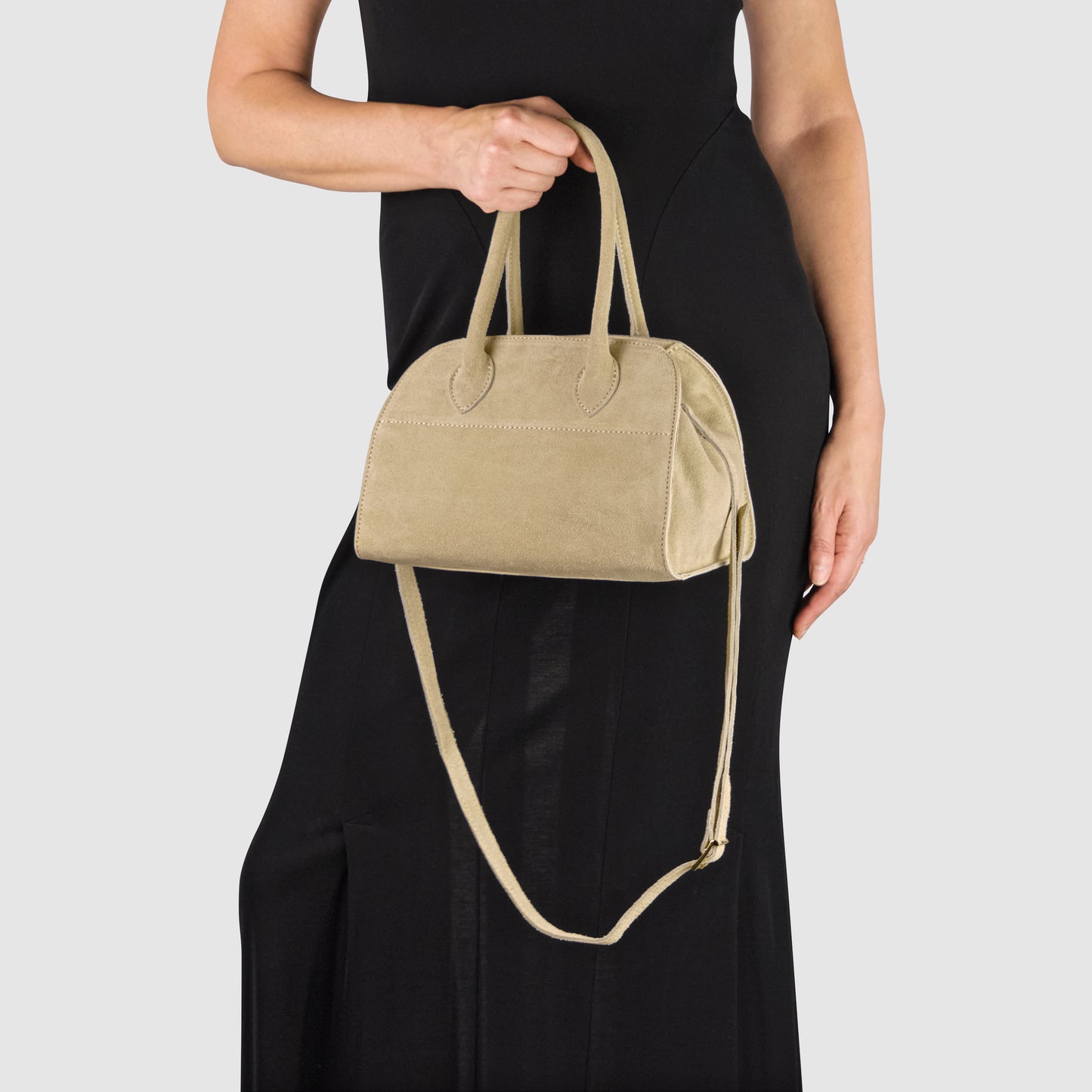 A person in a black dress holds a handmade Italian handbag by both handles, its long strap hanging loosely. The beige bag’s genuine suede finish stands out against the plain light gray background.