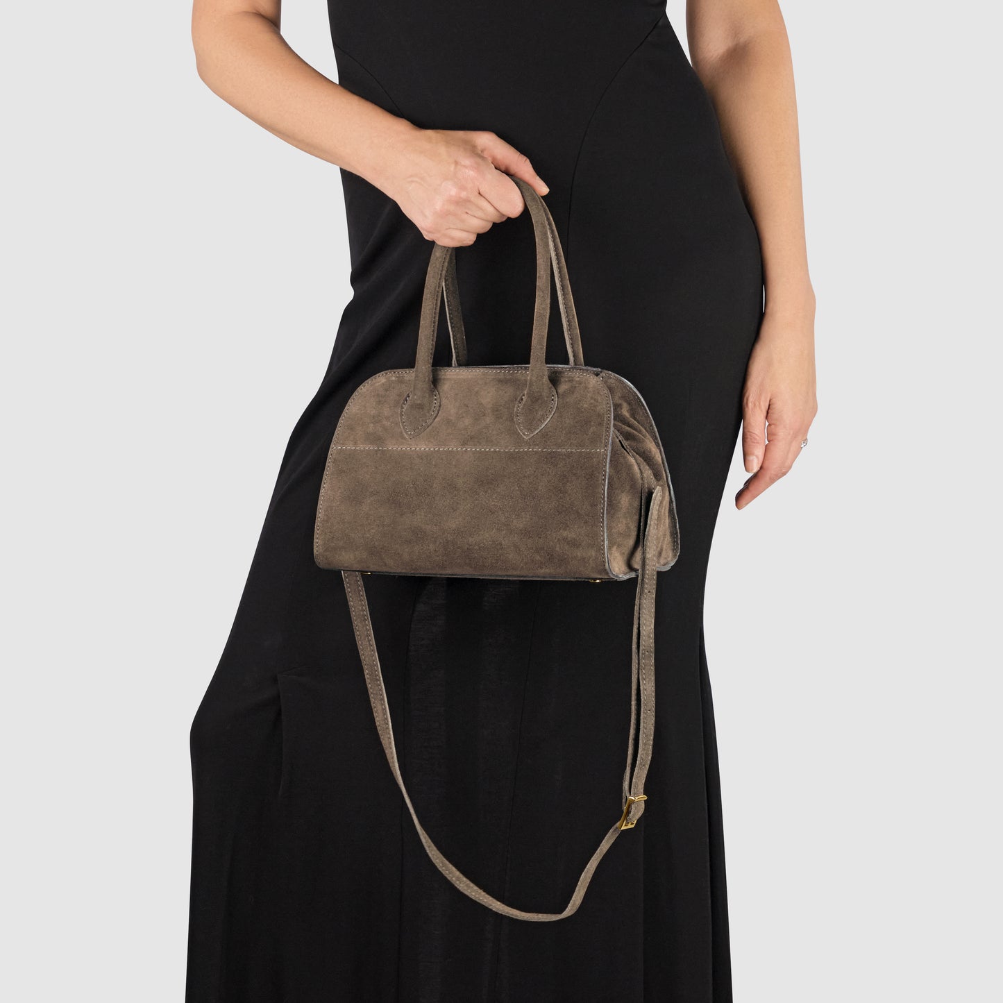 A person in a sleeveless black dress holds a luxury Italian handbag, handmade in Italy from taupe genuine suede, with top handles and a long shoulder strap against a plain light background.