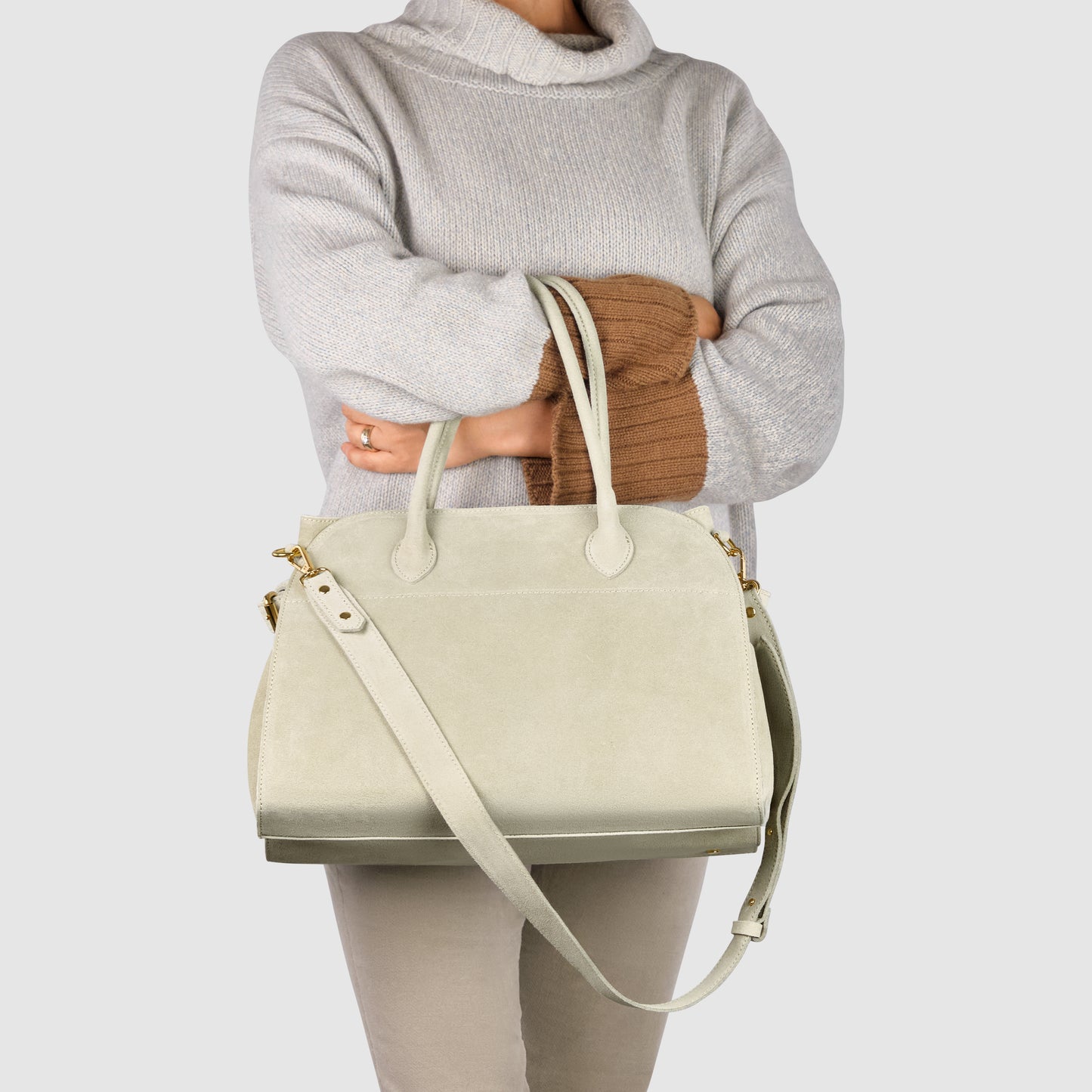 A person wearing a light gray sweater and beige pants holds a large, handcrafted leather purse with a shoulder strap and gold hardware against a plain background.