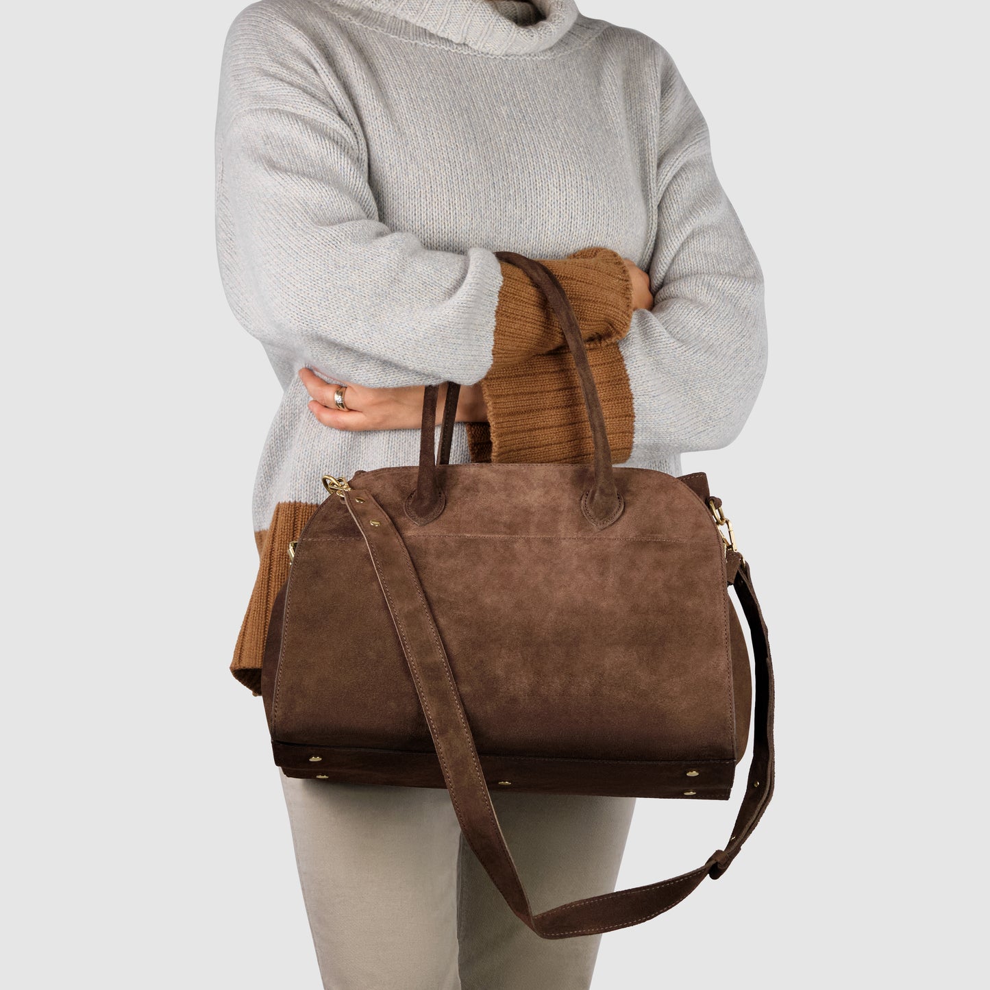 A person wearing a light gray sweater and beige pants holds a large handcrafted Italian suede doctor's bag purse with handles and a shoulder strap against a plain light background.