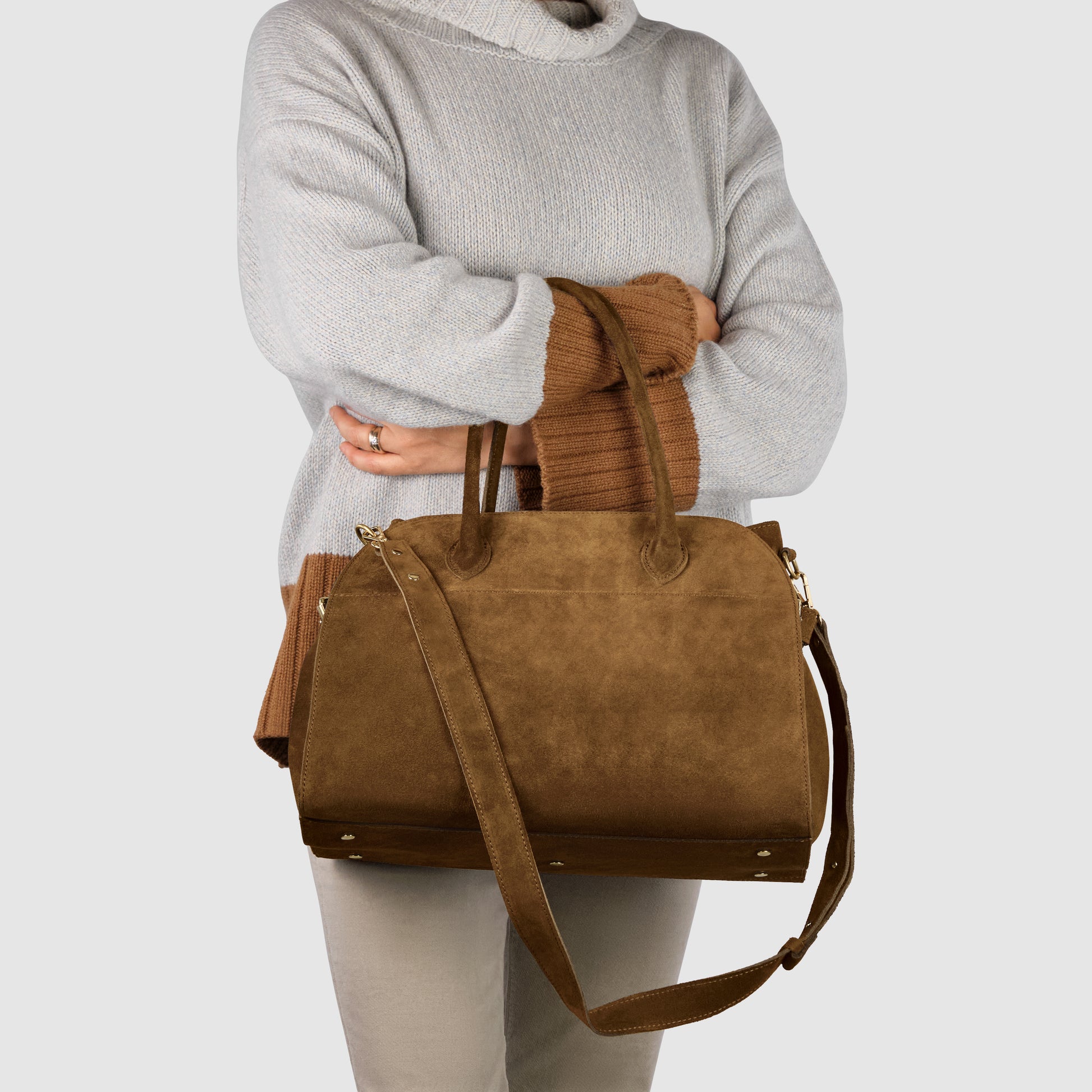 A person in a light gray sweater holds a large Italian suede handbag with both handles and a long shoulder strap, standing against a plain light background.