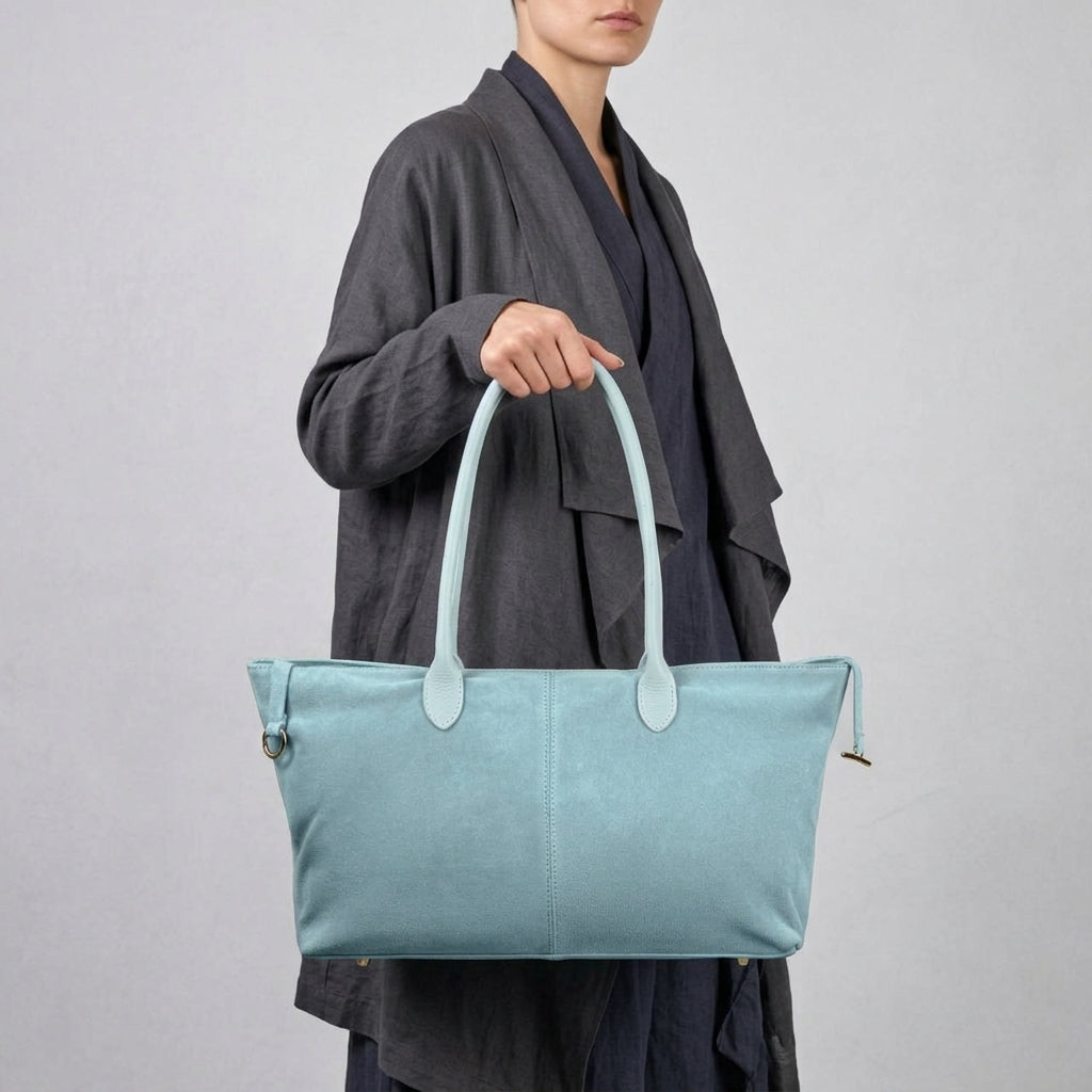 A person wearing a dark gray, draped outfit holds a large, light blue Italian suede tote bag with two handles against a plain light gray background.