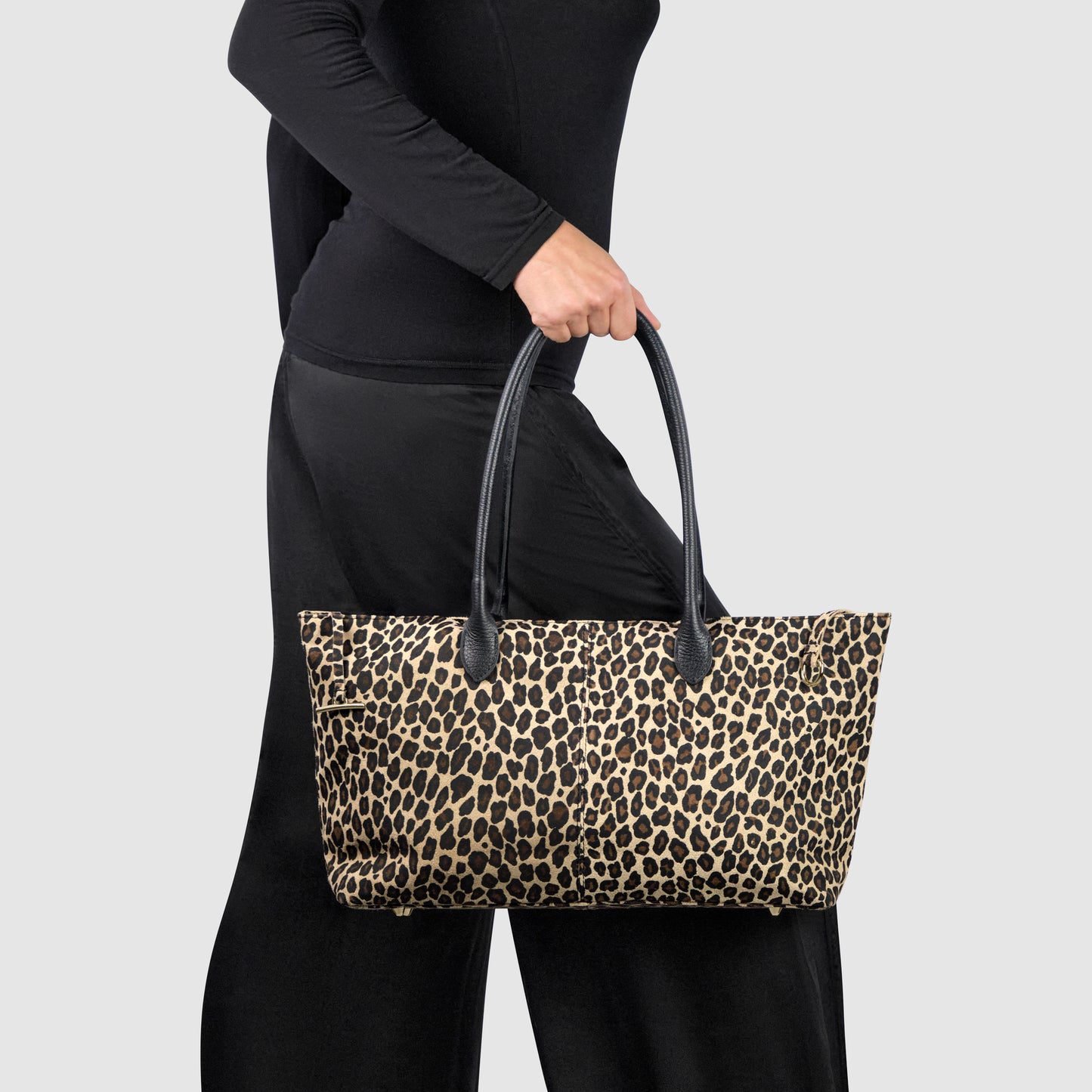 A person dressed in black holds an Italian-crafted tote with a leopard print pattern and black handles against a plain light gray background.