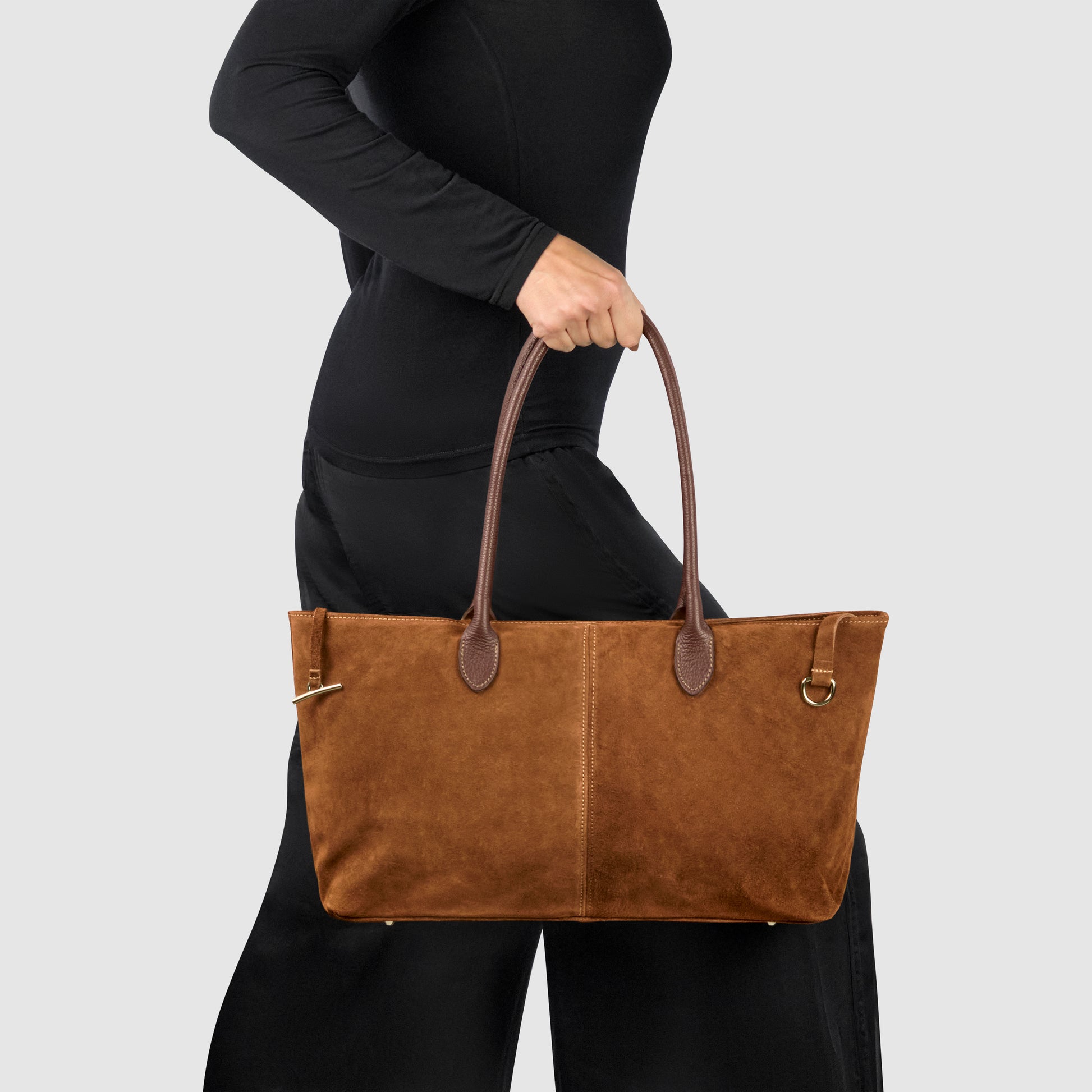 A person in black clothing holds an Italian-crafted suede tote bag with dark brown handles against a plain light gray background.