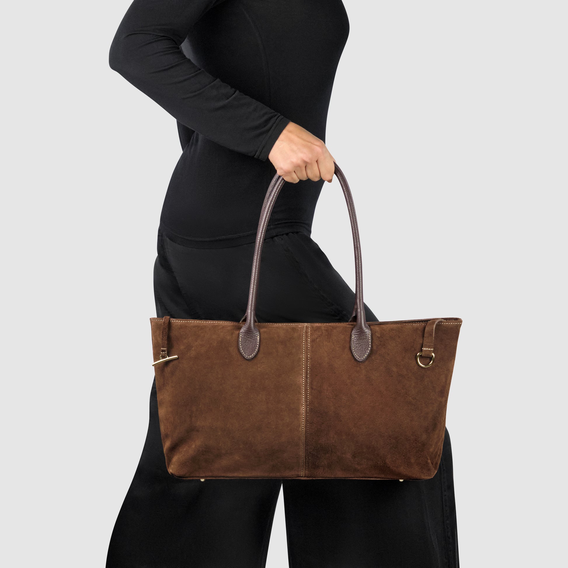 A person dressed in black carries an Italian suede tote bag with dark genuine leather handles against a plain light gray background.