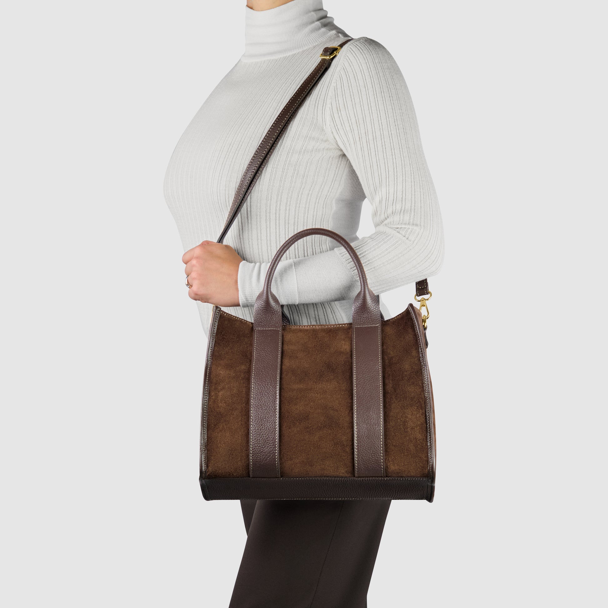 A person wearing a white ribbed turtleneck and dark pants holds a handmade Italian suede bag with dark leather straps and a matching crossbody shoulder strap against a plain light background.