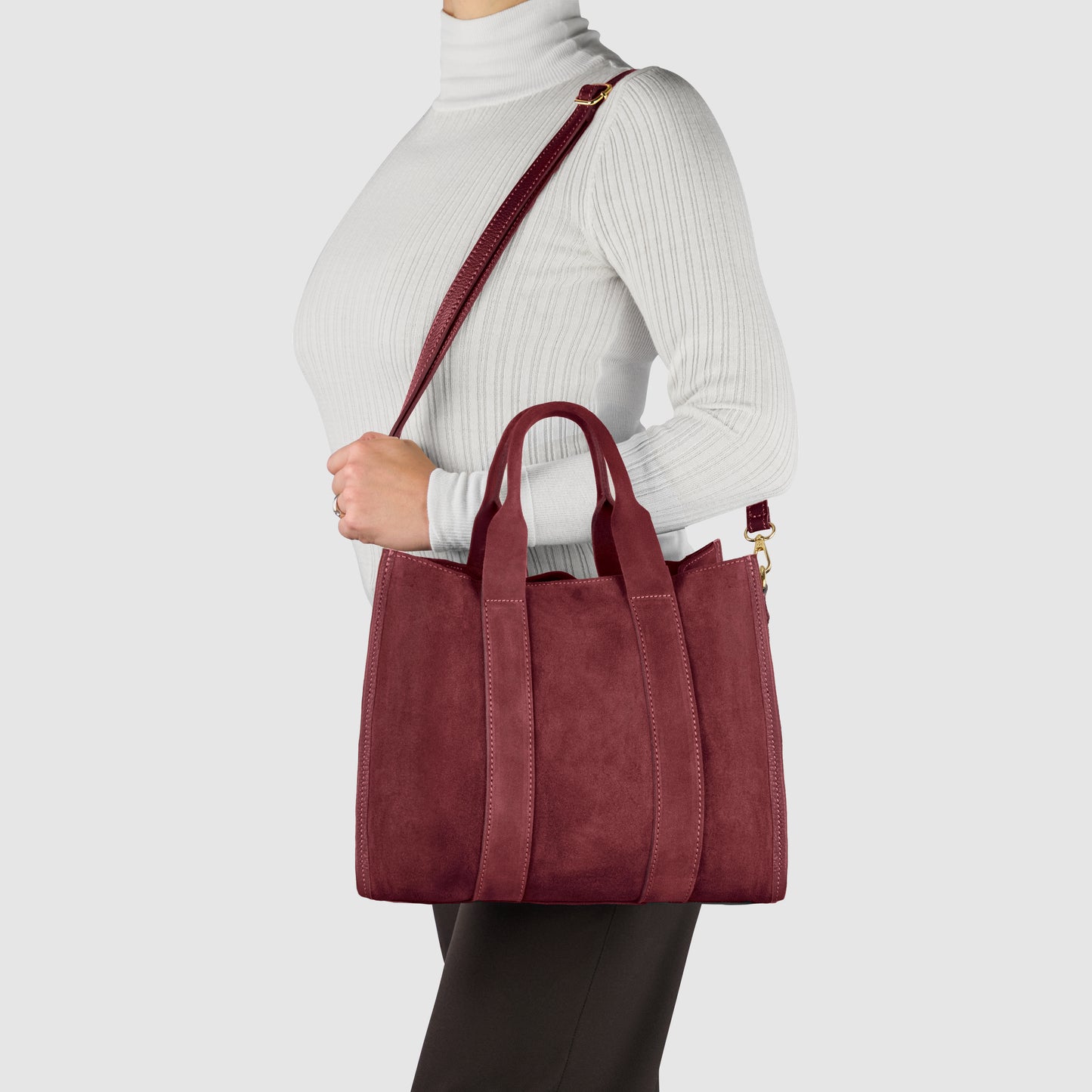 A person wearing a white turtleneck and black pants holds a burgundy suede carryall handmade in Italy, featuring top handles and a shoulder strap, against a plain light background.