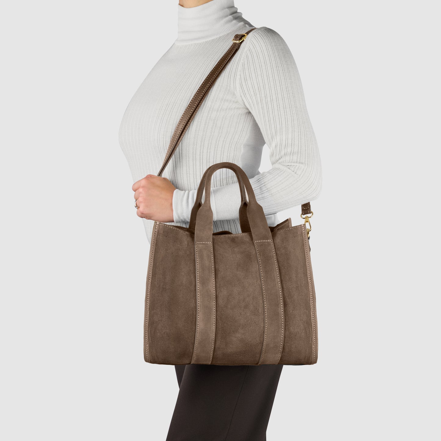 A person wearing a white turtleneck and black pants carries a brown suede carryall with two short handles and a long shoulder strap against a plain light background.