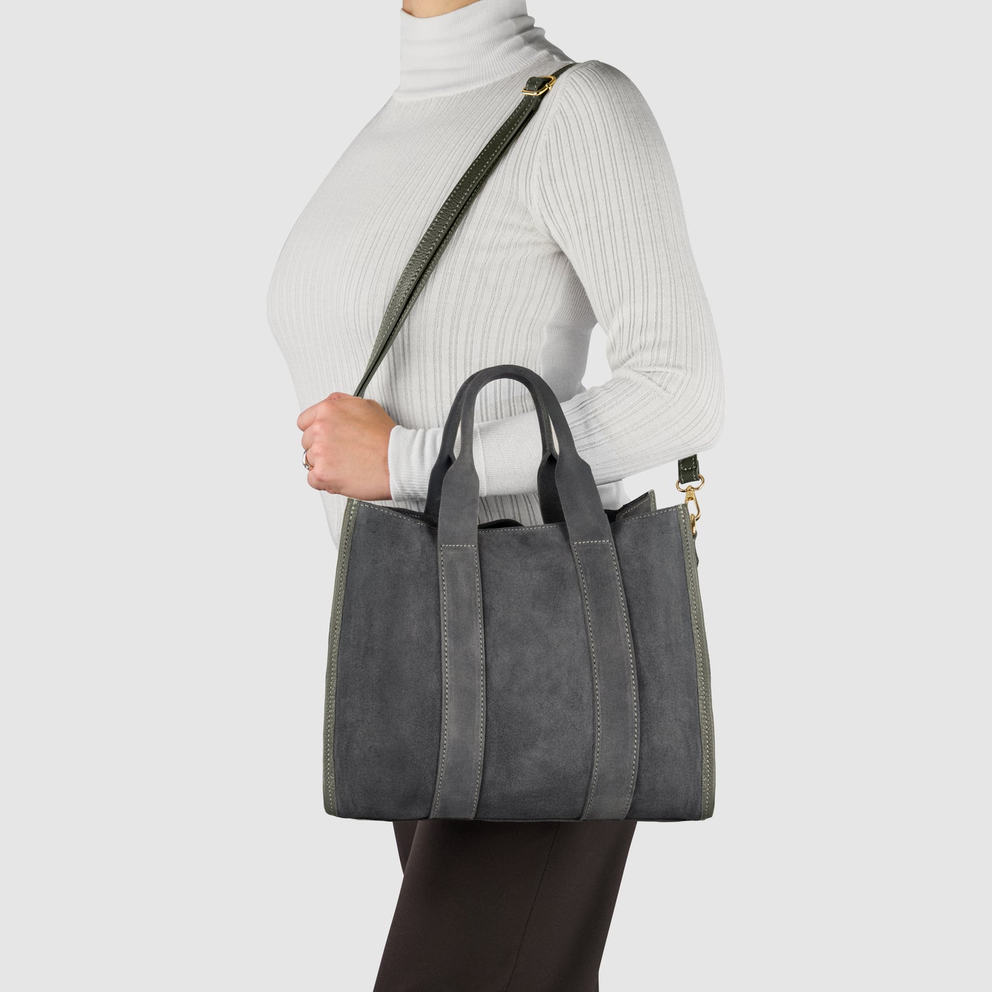 A person wearing a white turtleneck and black pants carries an Italian suede bag with short handles and a long strap over their shoulder against a plain light background.