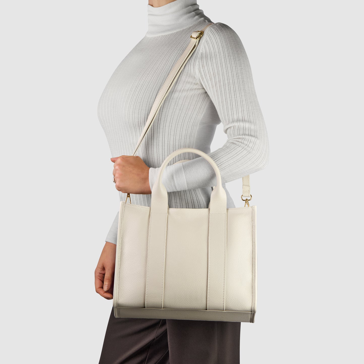 A person wearing a white turtleneck and dark pants holds a cream-colored handcrafted handbag with short handles and a long shoulder strap against a plain light background.