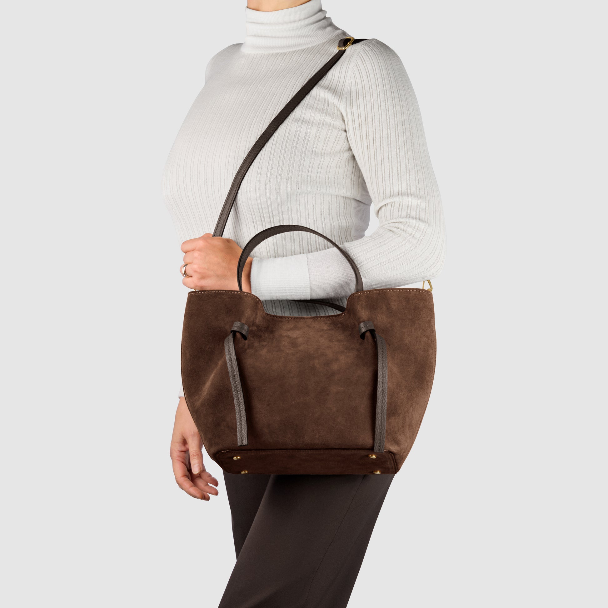 A person in a white ribbed turtleneck and dark pants carries a handmade Italian bag with top handles and a shoulder strap against a plain gray background.