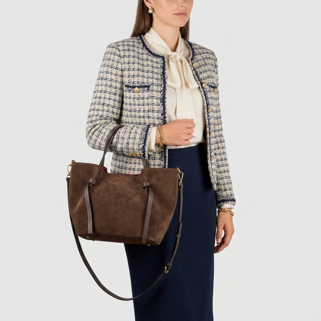 A woman dressed in a checked blazer, white blouse, and navy skirt is holding an Italian suede bag with both handles and a shoulder strap against a plain background.