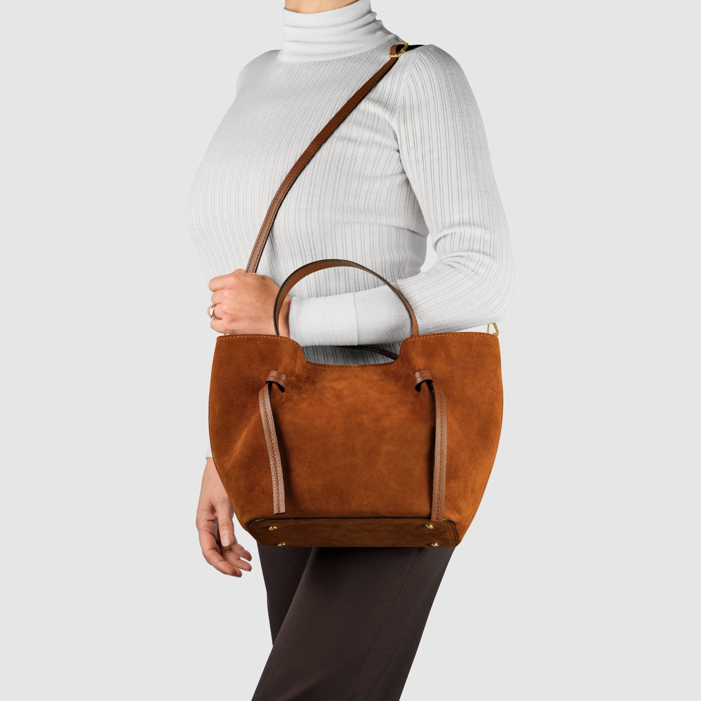 A person wearing a white ribbed turtleneck and dark pants carries a brown suede tote, handmade in Italy, with two handles and a shoulder strap against a plain light gray background.
