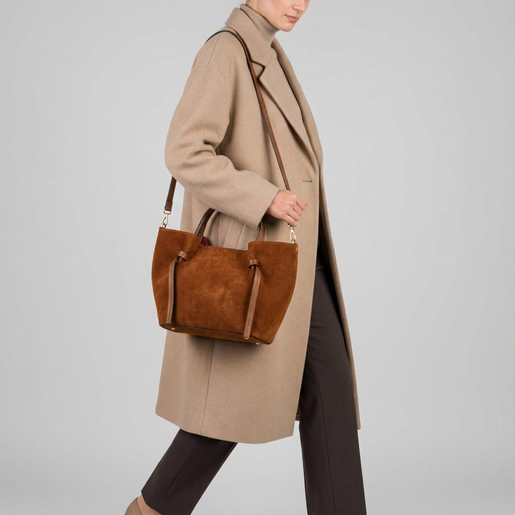 A person wearing a long beige coat and brown pants carries an Italian suede bag with shoulder straps. The background is plain light gray.