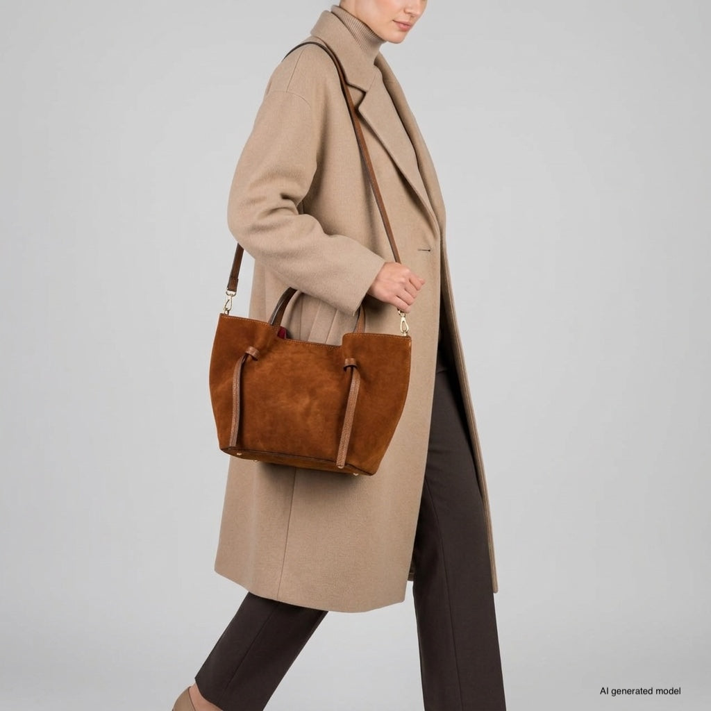 A person wearing a beige coat and brown pants carries a handmade Florentine bag with shoulder straps. The Italian suede bag stands out against the plain gray background.