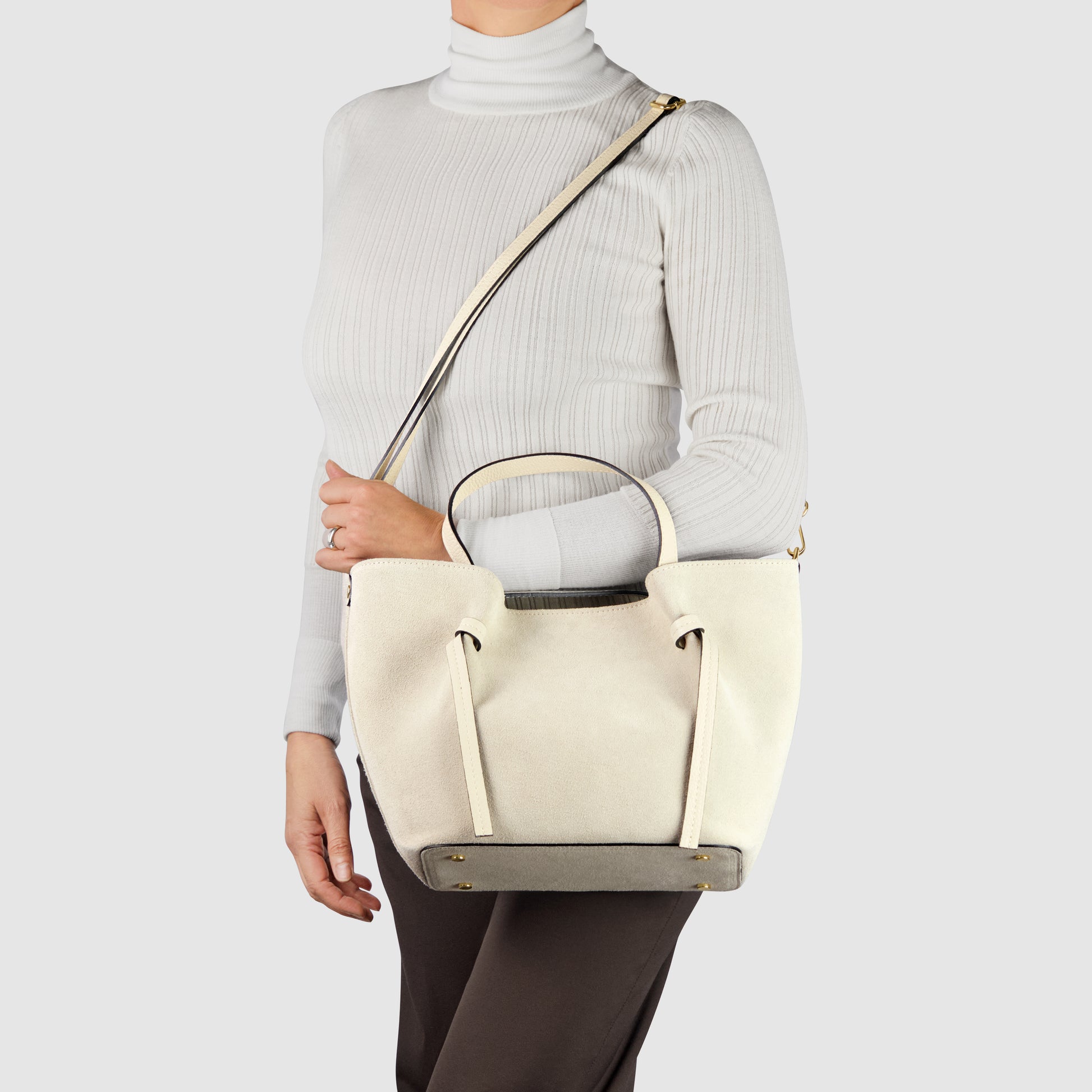A woman wearing a white turtleneck and dark pants holds a luxury tote with short handles and a long shoulder strap, showcasing Florentine leatherwork against a plain light background.