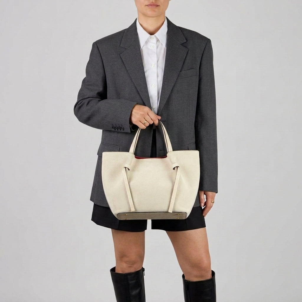 A person wearing a gray blazer, white shirt, black shorts, and black knee-high boots holds a handmade Florentine bag—perfect as an everyday carryall—against a plain light background. Their face is not fully visible.