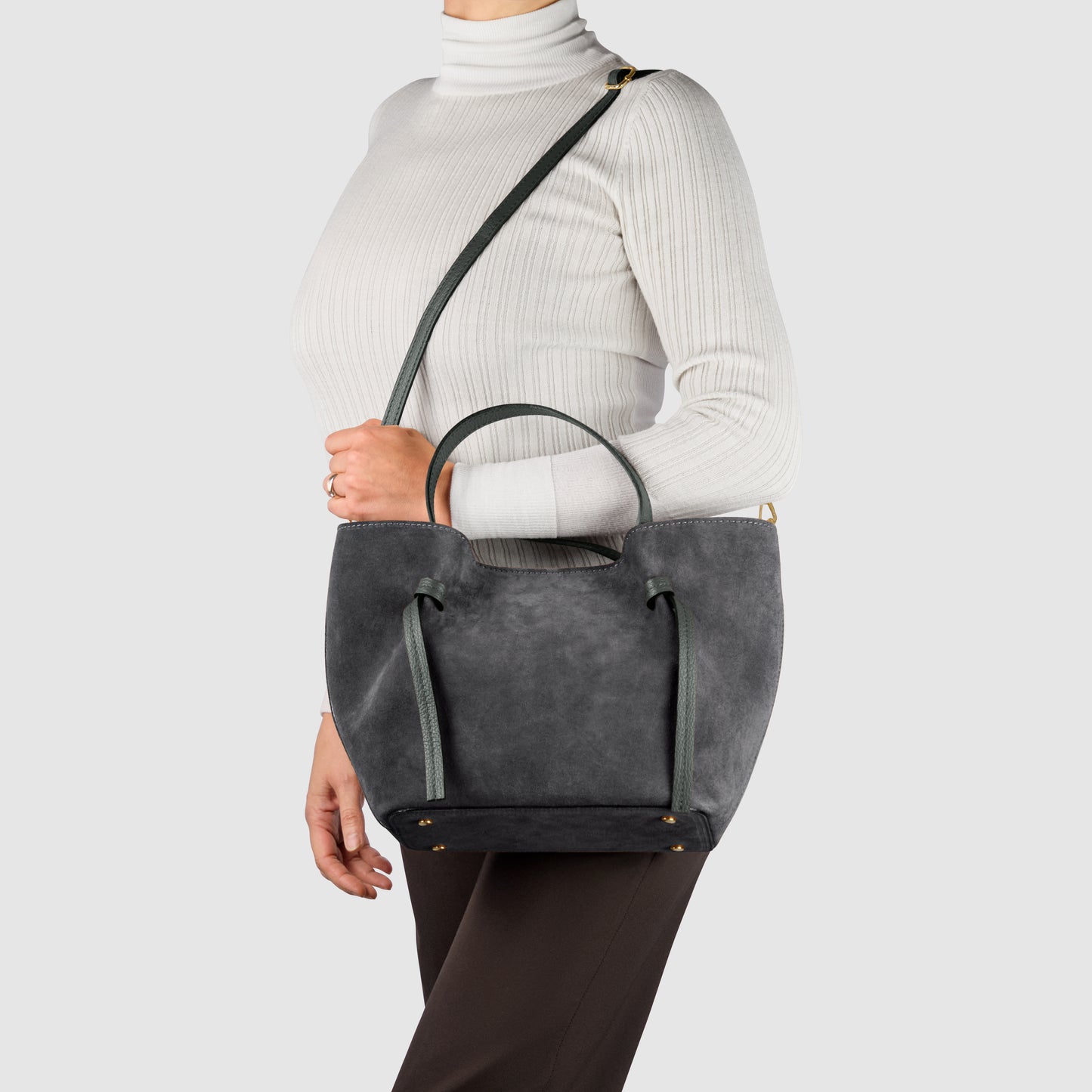 A person wearing a white turtleneck and dark pants is holding an Italian suede bag—showcasing classic Florentine leatherwork—with both a shoulder strap and short handles against a plain light background.