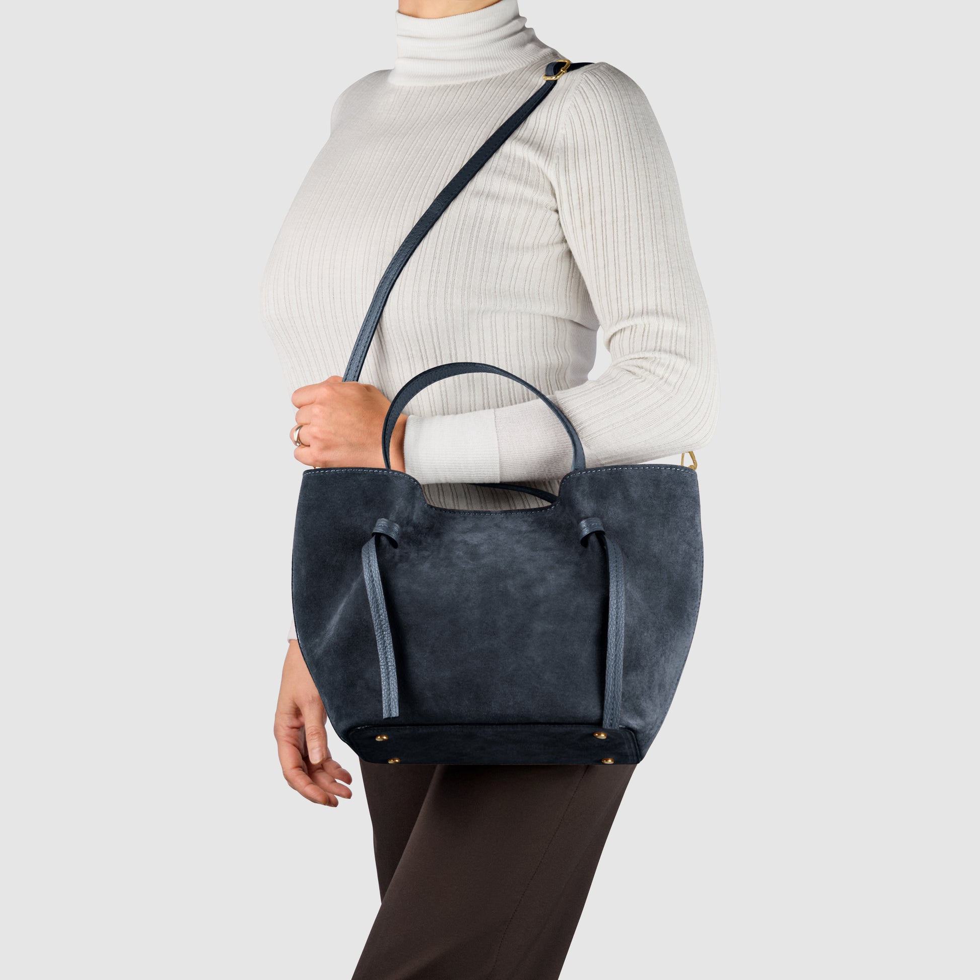 A person wearing a white ribbed turtleneck and brown pants carries a dark blue handmade Italian bag with both short handles and a shoulder strap against a gray background.