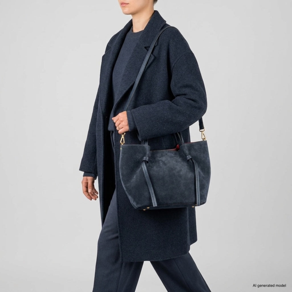 A person wearing a dark navy coat and pants carries a large, handmade Florentine bag in dark blue suede over their shoulder. The background is plain and light grey.