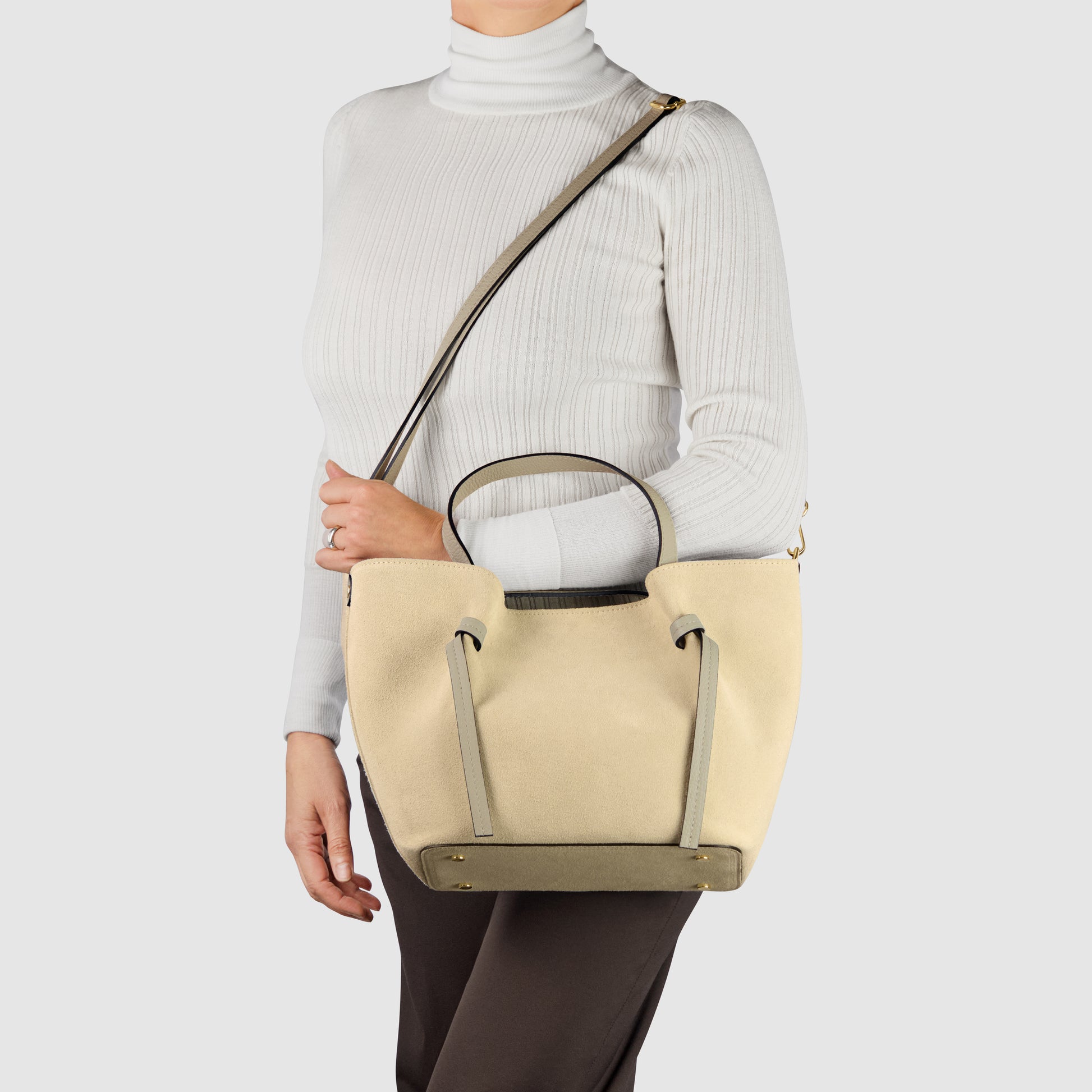 A person wearing a white turtleneck and dark pants holds a handmade Italian bag with short handles and a long shoulder strap, showcasing classic Florentine leatherwork against a plain light background.