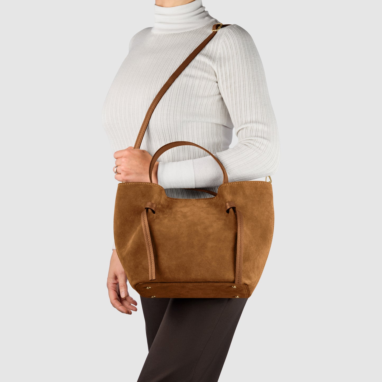 A person wearing a white ribbed turtleneck and dark pants carries a handmade Italian bag in brown suede with both short handles and a long shoulder strap, seen against a plain light gray background.