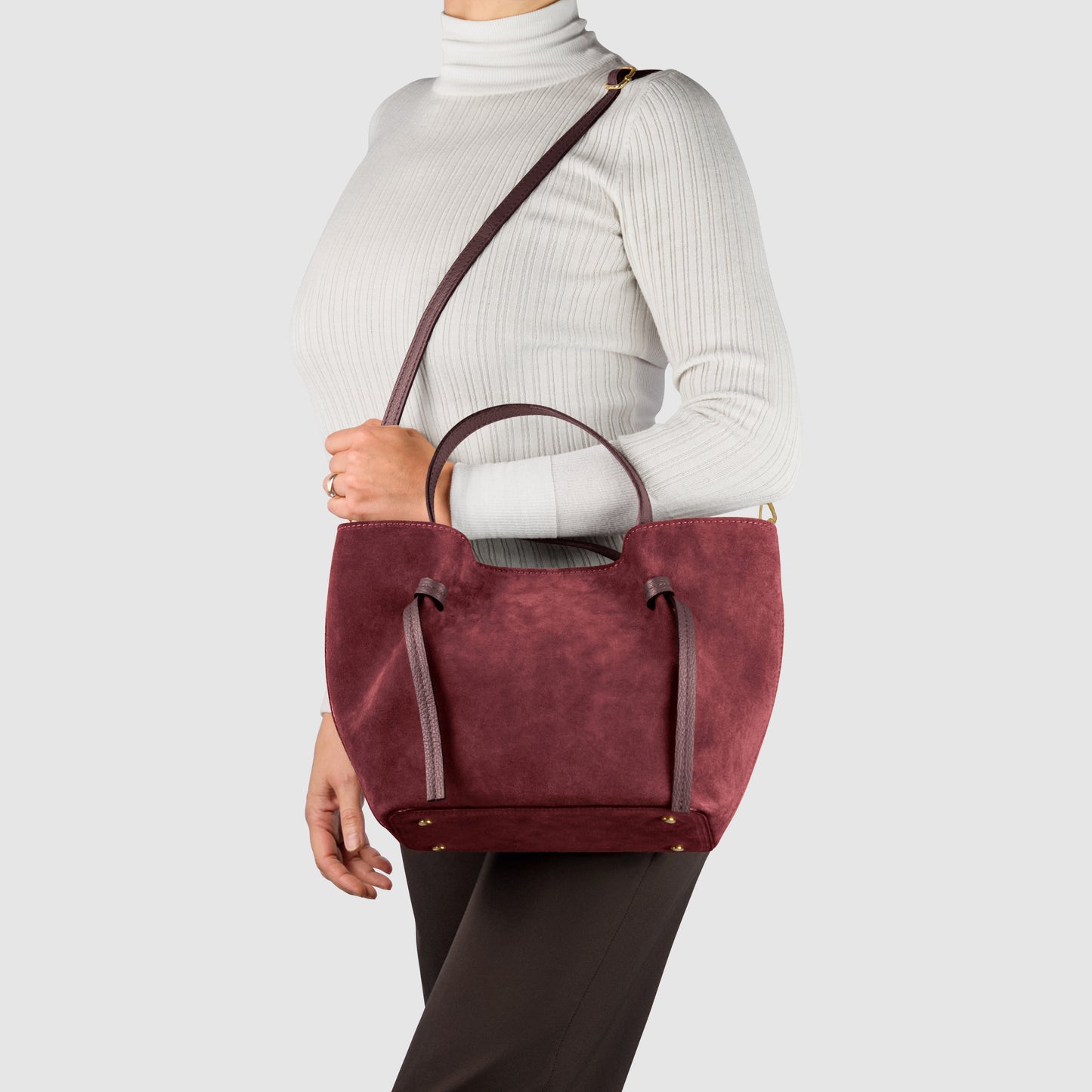 A woman wearing a white ribbed turtleneck and dark pants carries a handmade Italian bag—a burgundy suede tote with top handles and a shoulder strap—against a plain light background.