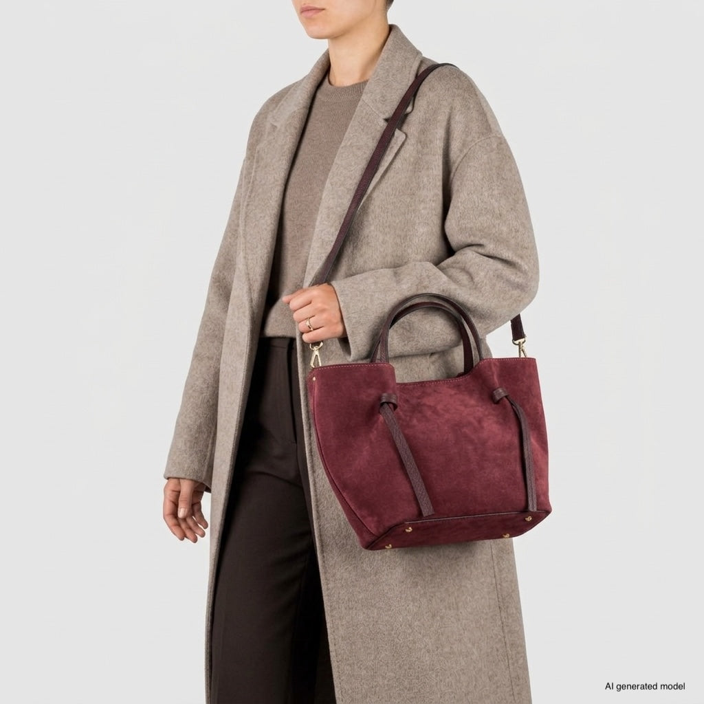 A person wearing a long beige coat over a taupe top and dark trousers holds an Italian suede everyday carryall with top handles and a shoulder strap against a plain light background.