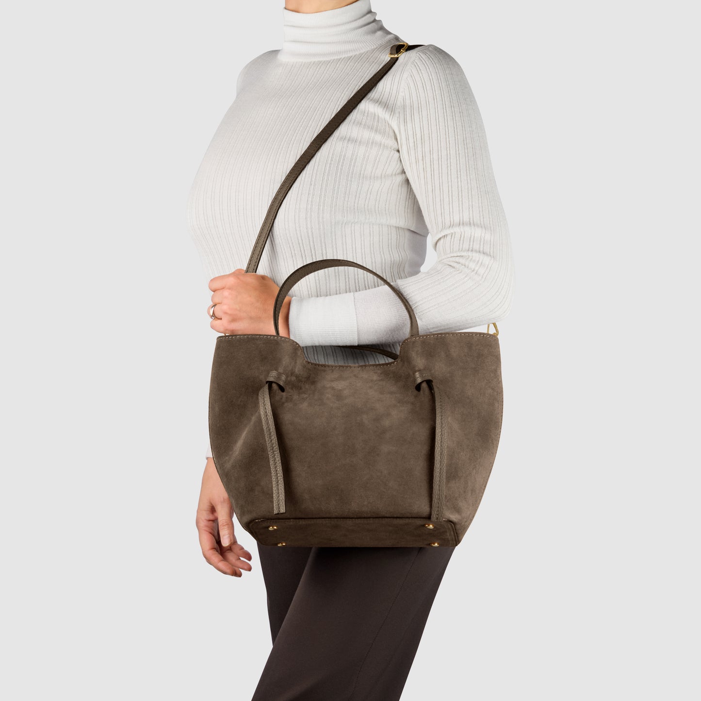 A woman wearing a white turtleneck and dark pants carries a handmade Italian bag with top handles and a shoulder strap, crafted in brown suede, against a plain light background.