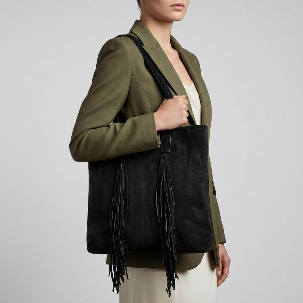 A woman in an olive green blazer and cream dress holds a large black suede handbag with long fringe tassels, styled as a boho bag. The background is plain and light gray.