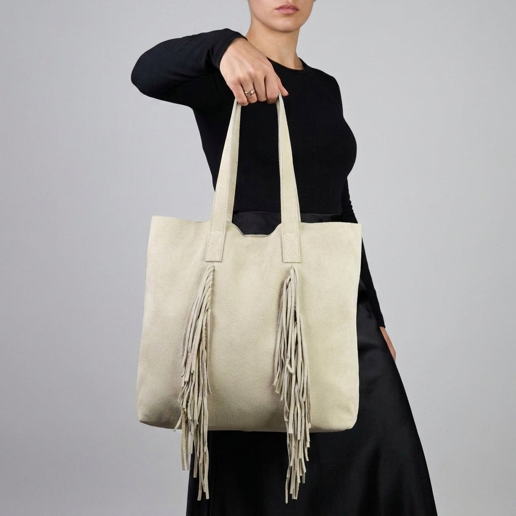 A person in a black long-sleeve top and skirt holds a beige boho bag with long fringe details on the front against a plain light gray background. The person's face is partially out of frame.