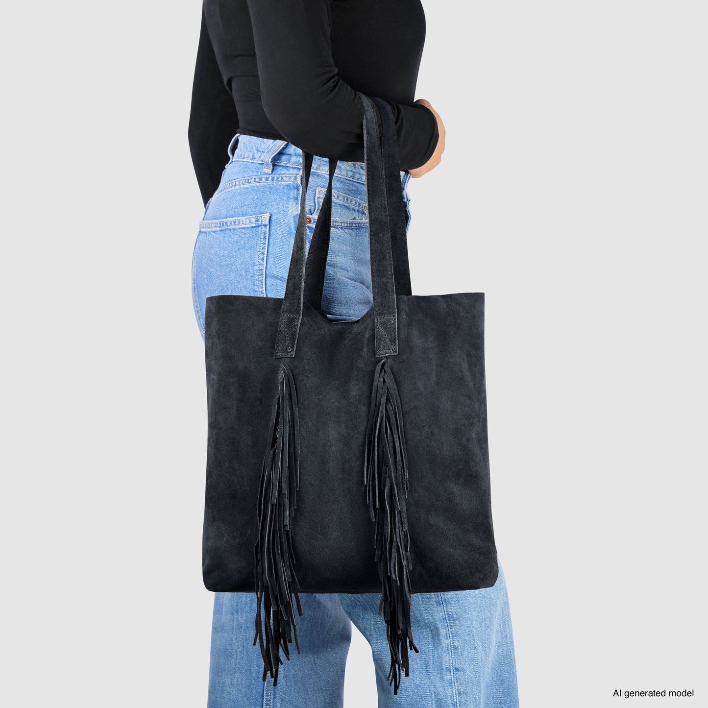 A person wearing a black long-sleeve top and blue jeans holds a large black suede handbag with long fringe detailing, draped over their shoulder against a plain light gray background.