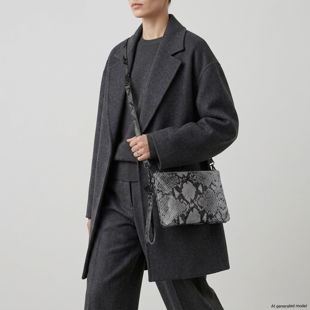 A person in a dark gray coat and pants holds a rectangular, python-embossed crossbody purse with a matching strap against a plain, light background. The genuine suede leather accessory is made in Italy; the person’s head is partially out of frame.