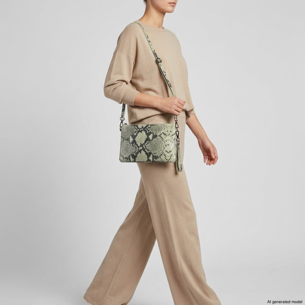 A person wearing a loose-fitting beige sweater and matching wide-leg pants carries a green and beige python-embossed crossbody purse. The background is plain light gray.