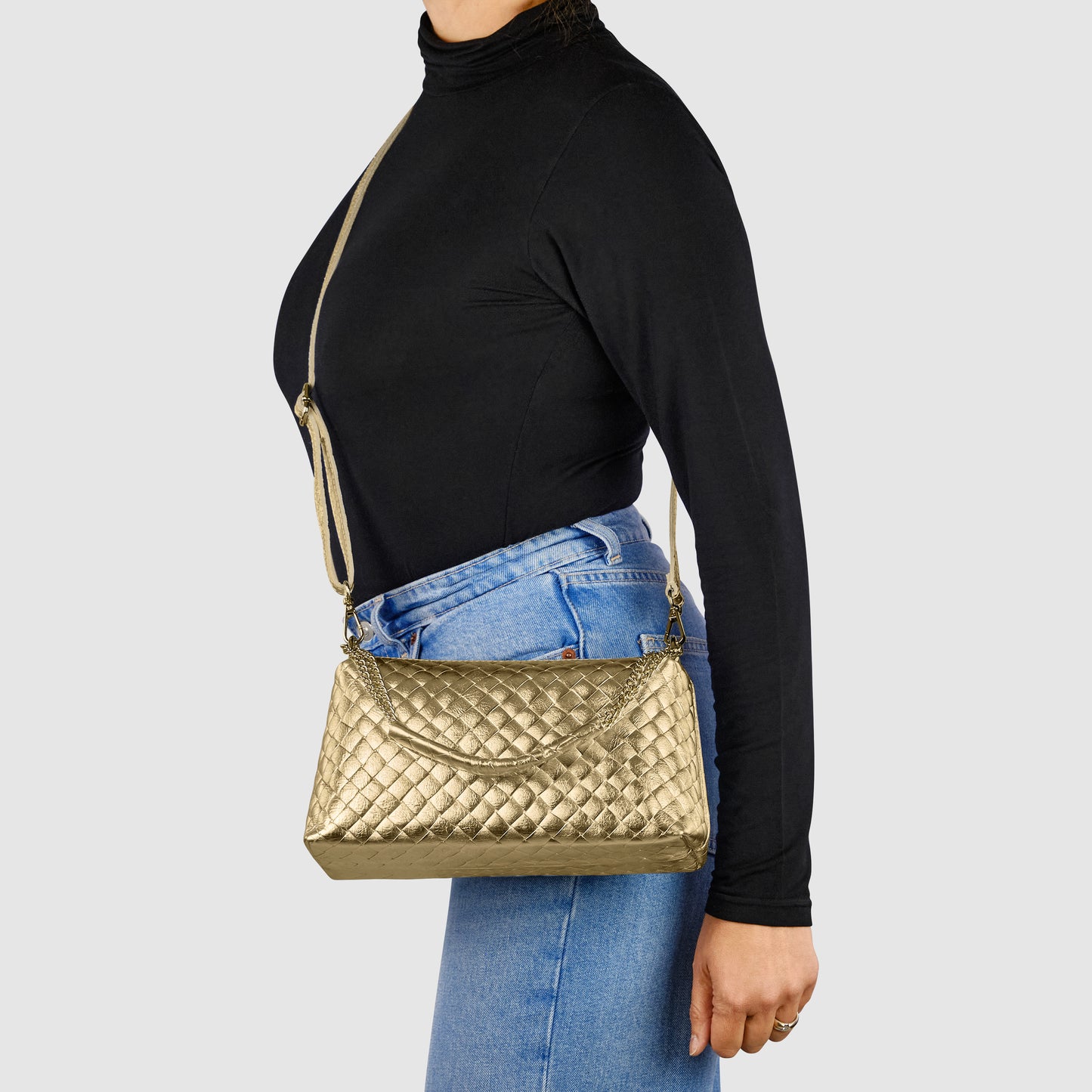 A person wearing a black turtleneck and blue jeans carries a gold quilted shoulder bag, reflecting exquisite Italian craftsmanship, with a long strap against a plain light gray background.