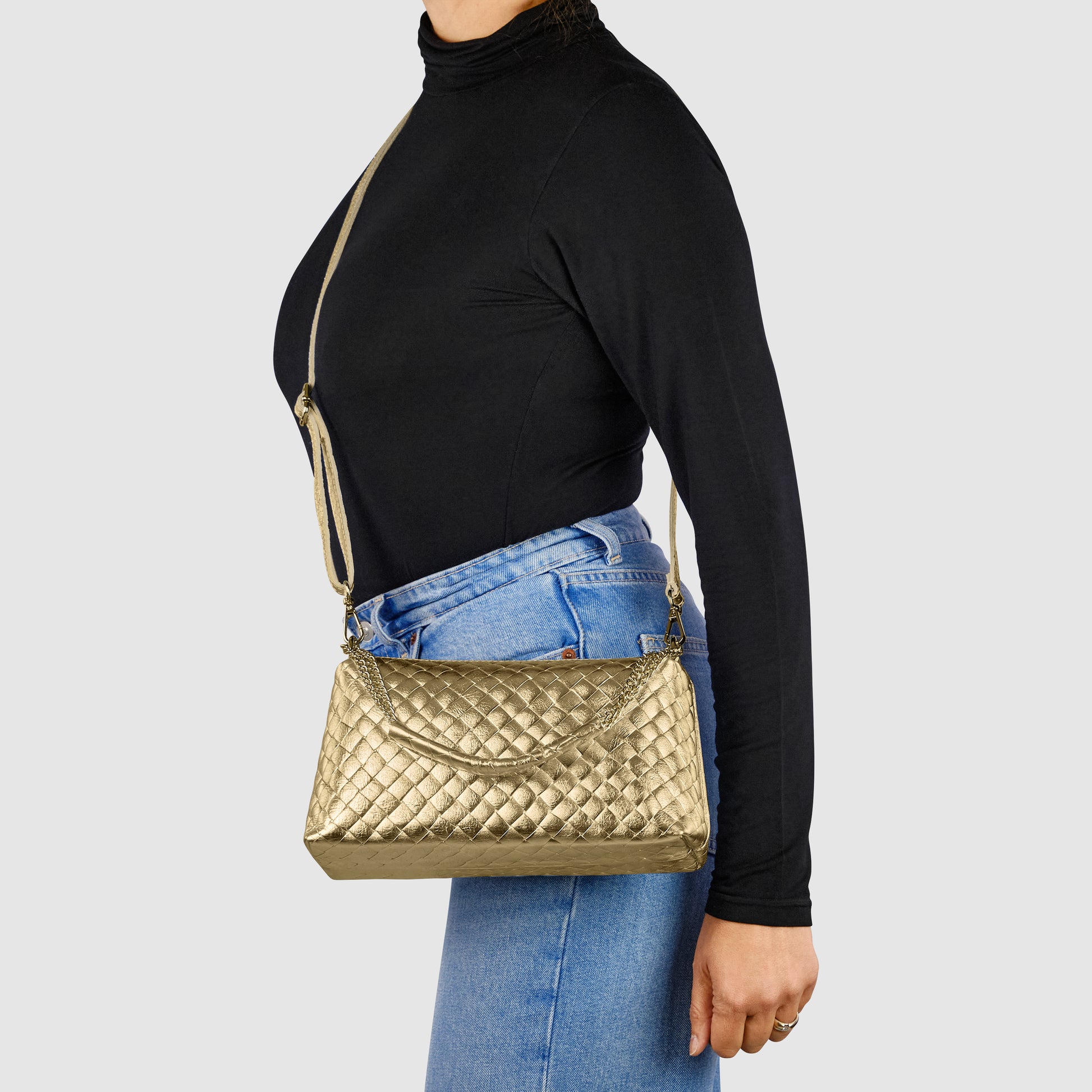 A person wearing a black turtleneck and blue jeans carries a gold quilted shoulder bag, reflecting exquisite Italian craftsmanship, with a long strap against a plain light gray background.