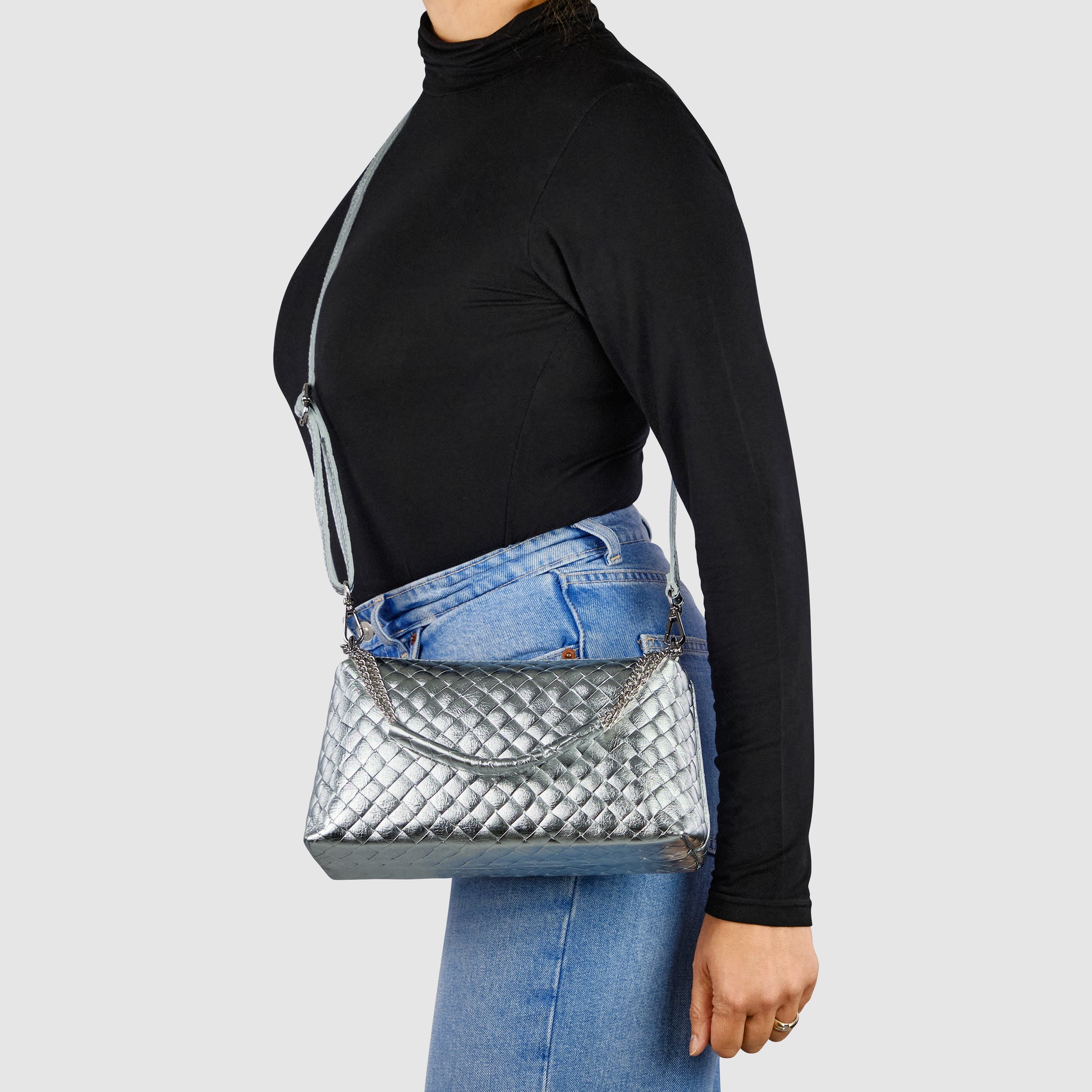 A person wearing a black turtleneck and blue jeans carries a silver suede leather miniature handbag with a top handle and shoulder strap, showcasing Italian craftsmanship against a plain light background.