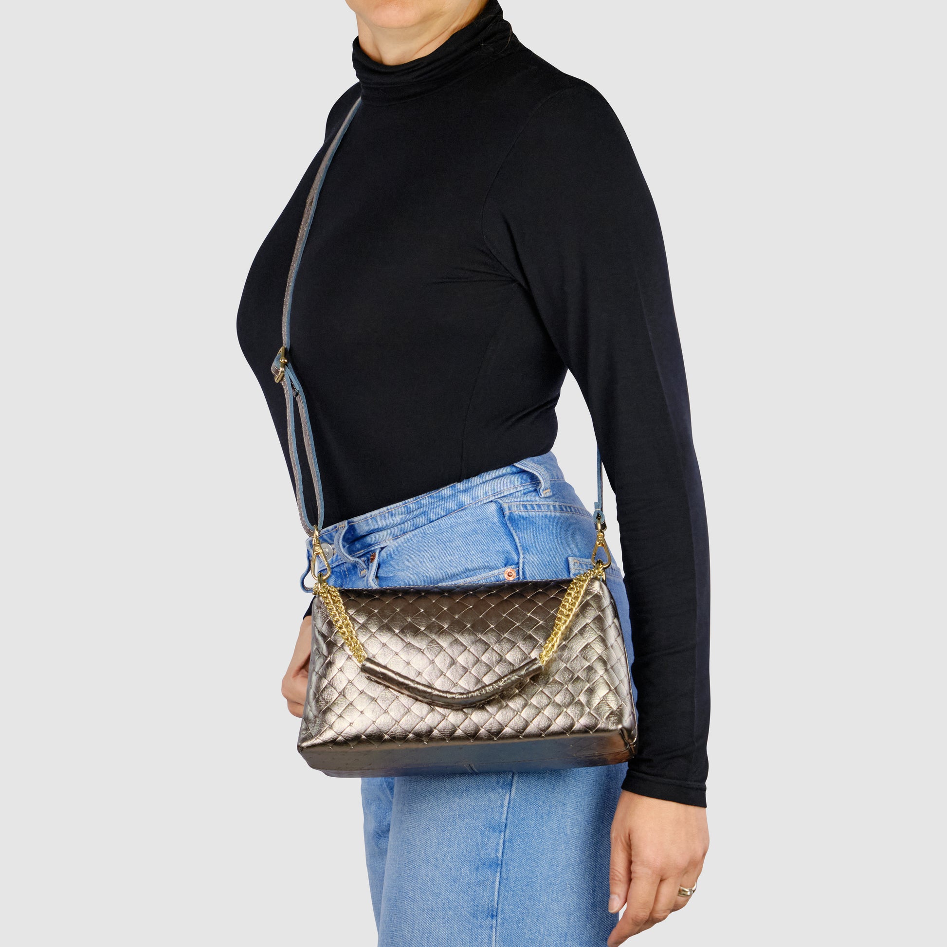 A person in a black turtleneck and blue jeans wears a metallic quilted miniature handbag with a chain strap, showcasing refined Italian craftsmanship against a plain light background.