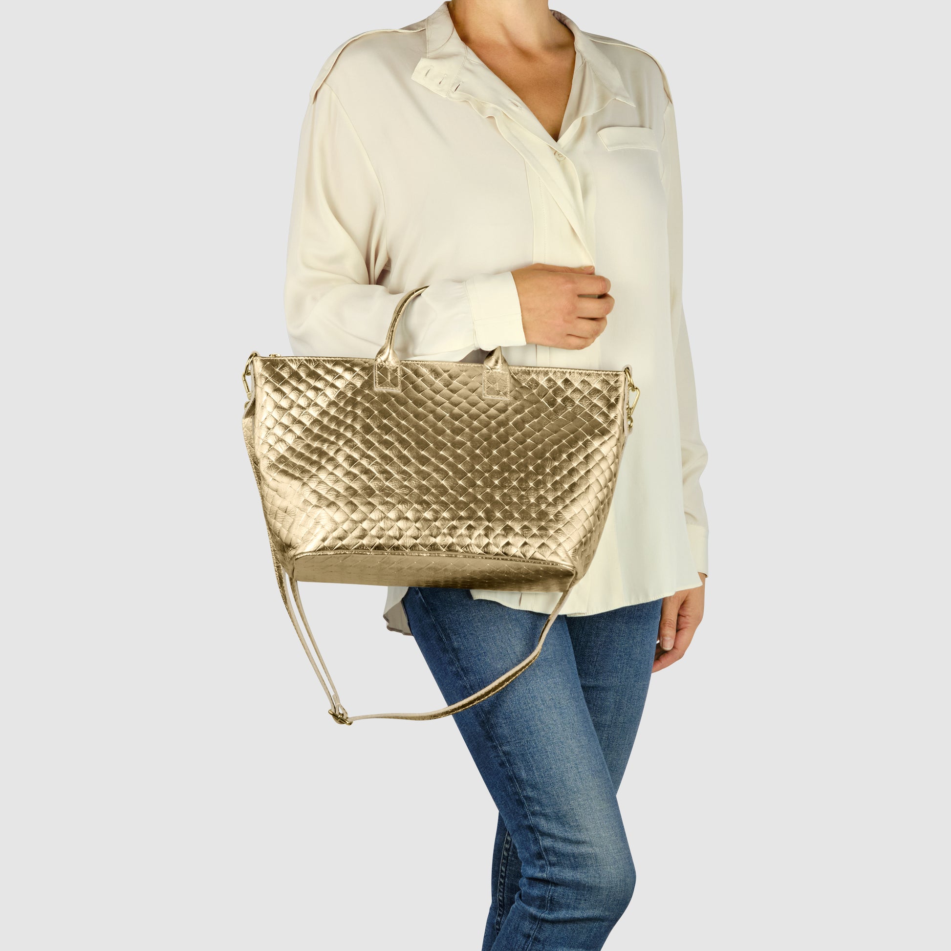 A woman wearing a cream-colored blouse and blue jeans holds a large, gold quilted handcrafted Italian handbag with both top handles and a shoulder strap against a plain light background.